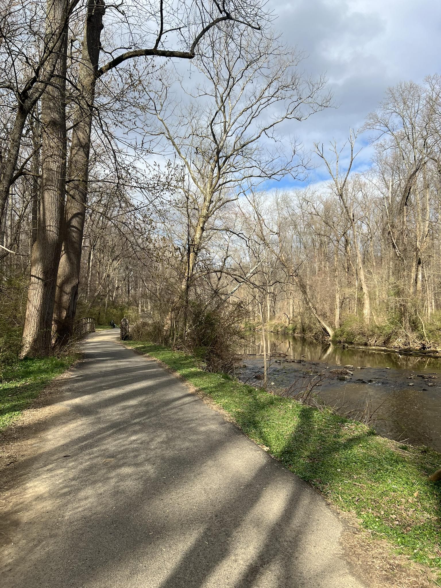 A paved path runs alongside a river in a wooded area under a cloudy sky. - Pennypack Trail Loop