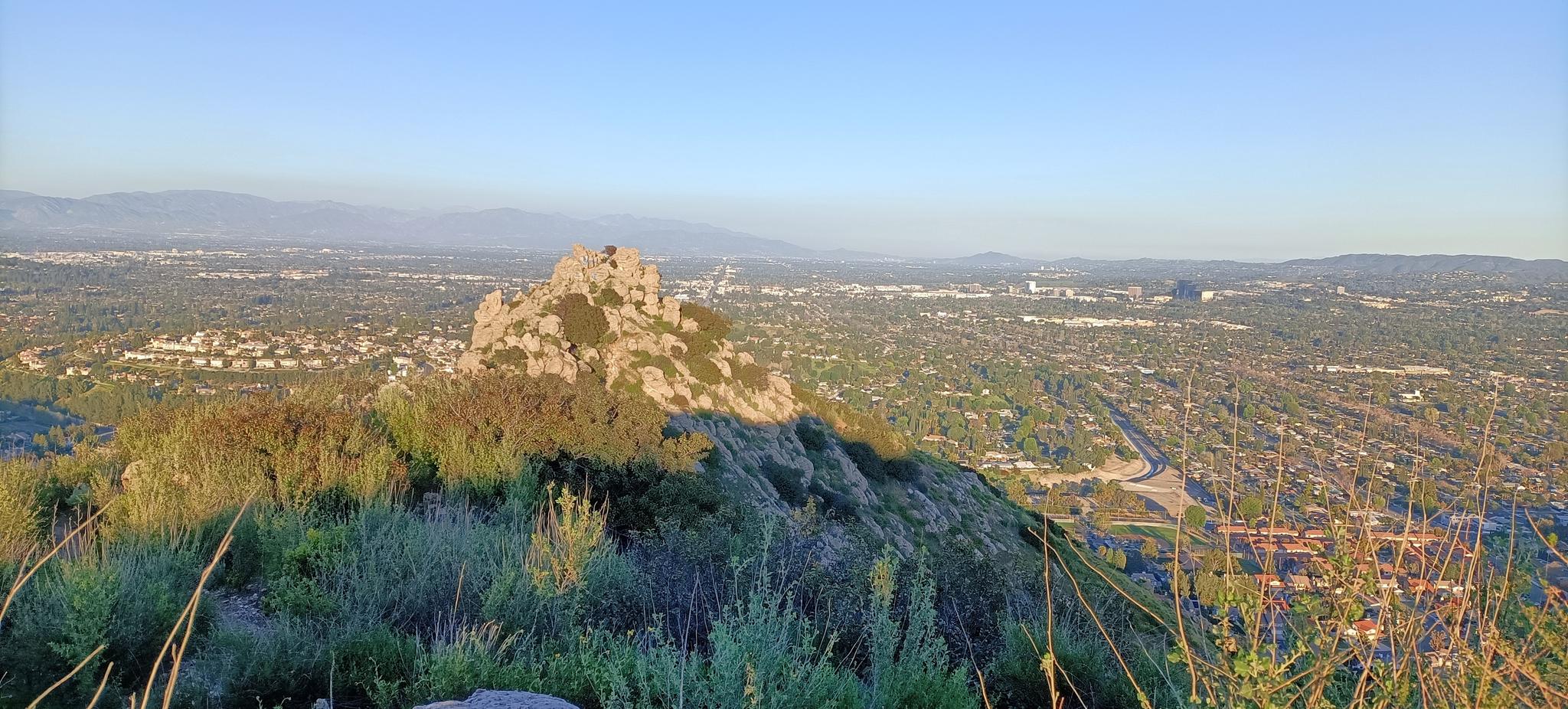 Rocky outcrop overlooking a sprawling city and distant mountains at sunset.