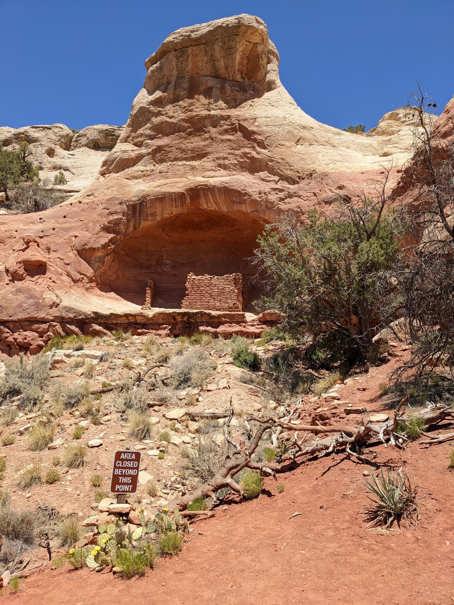 Ancient dwelling built into a red rock cave, with a sign on a dirt path.