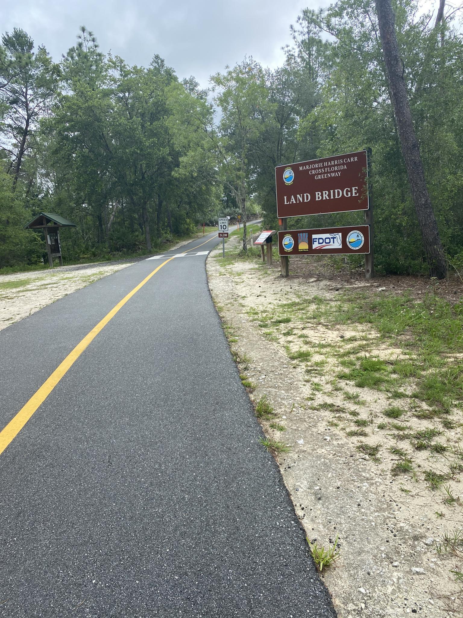 Paved path with a yellow line next to a sign, surrounded by trees.