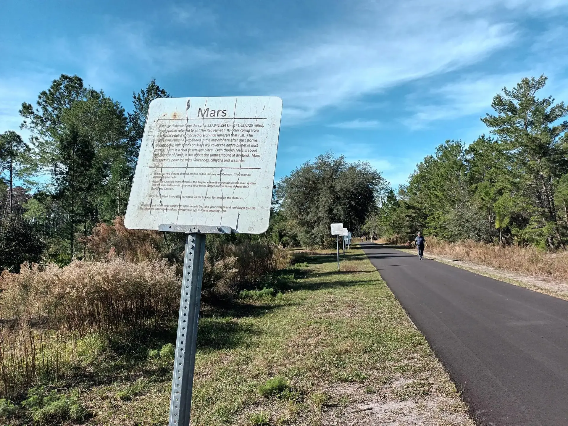 Informational sign next to a paved path with a person walking in sunlight.