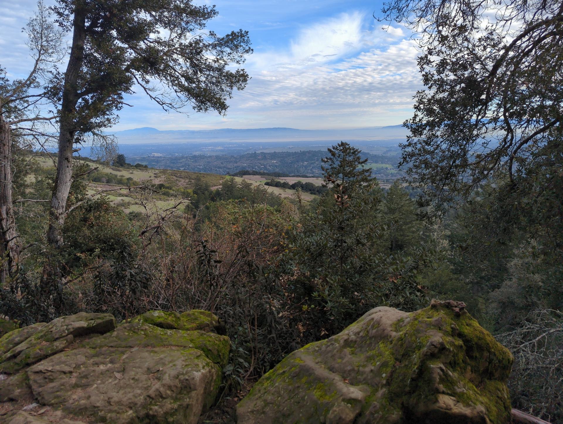 Mossy rocks and trees frame a distant valley under a cloudy sky.