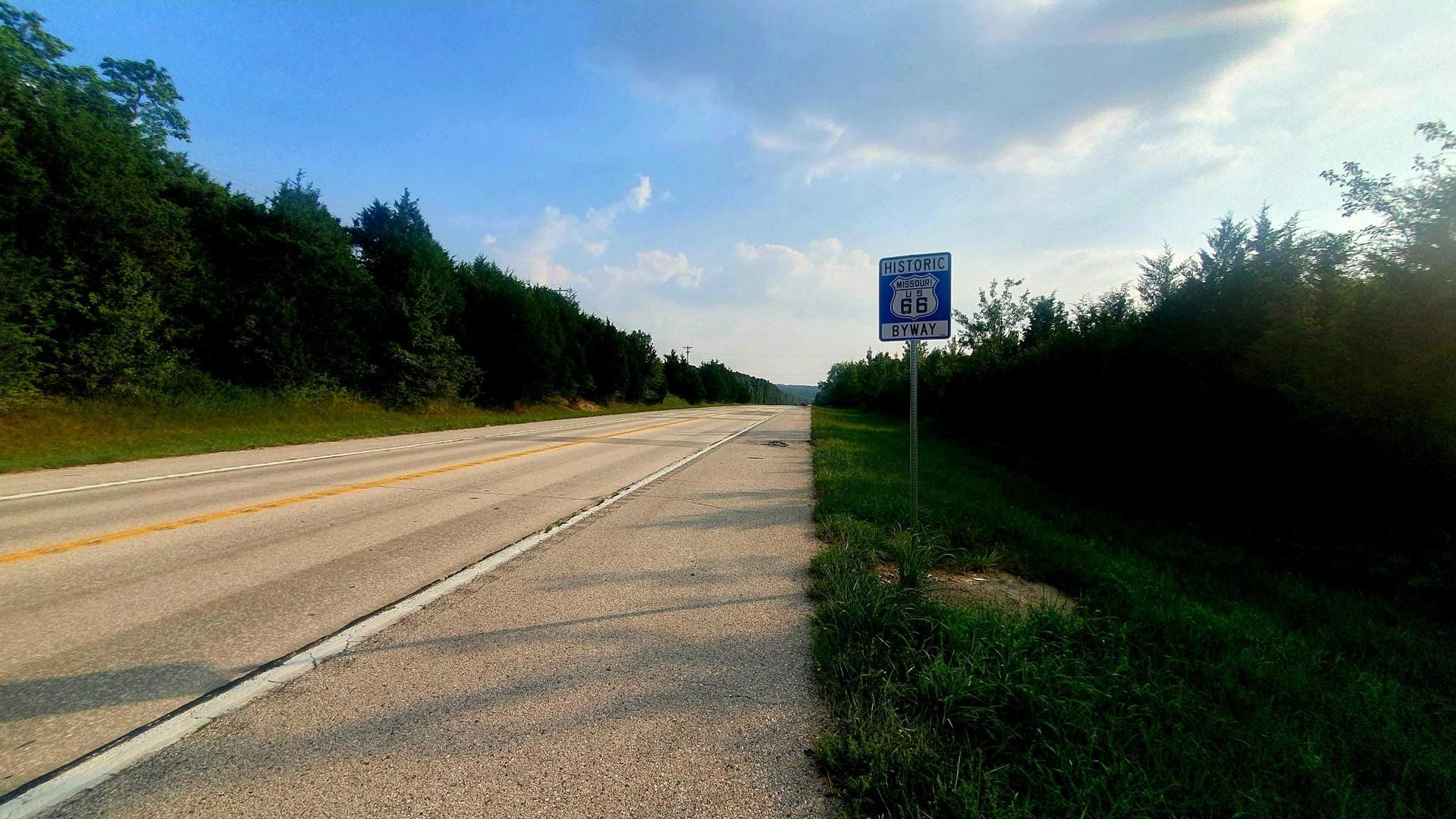 An empty road with a sign on the shoulder under a bright, cloudy sky.