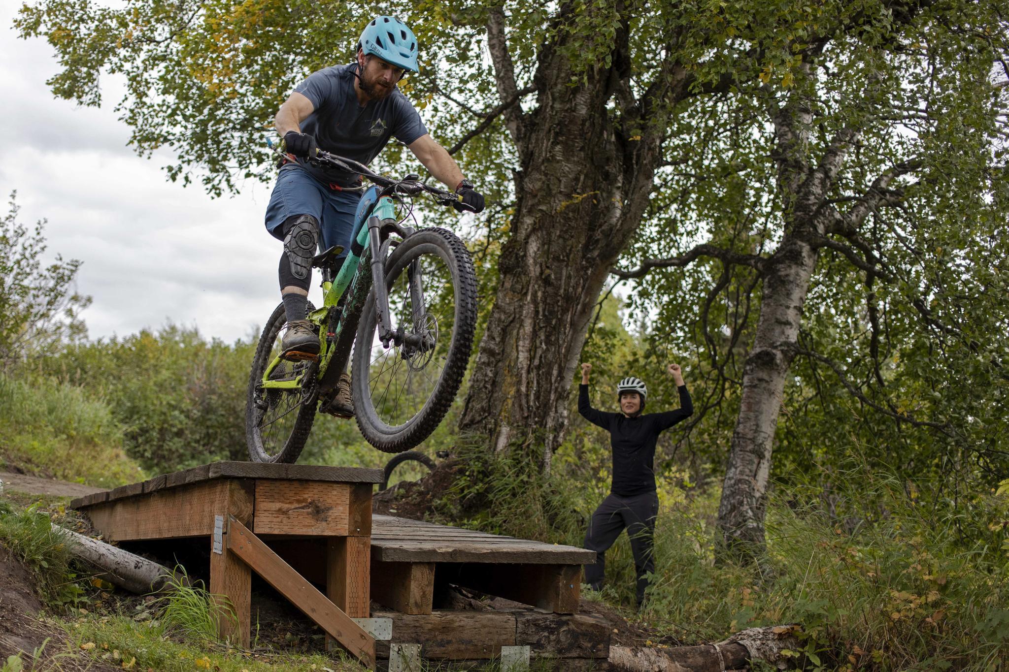 Man on mountain bike jumps off wooden ramp, person cheers, overcast day.