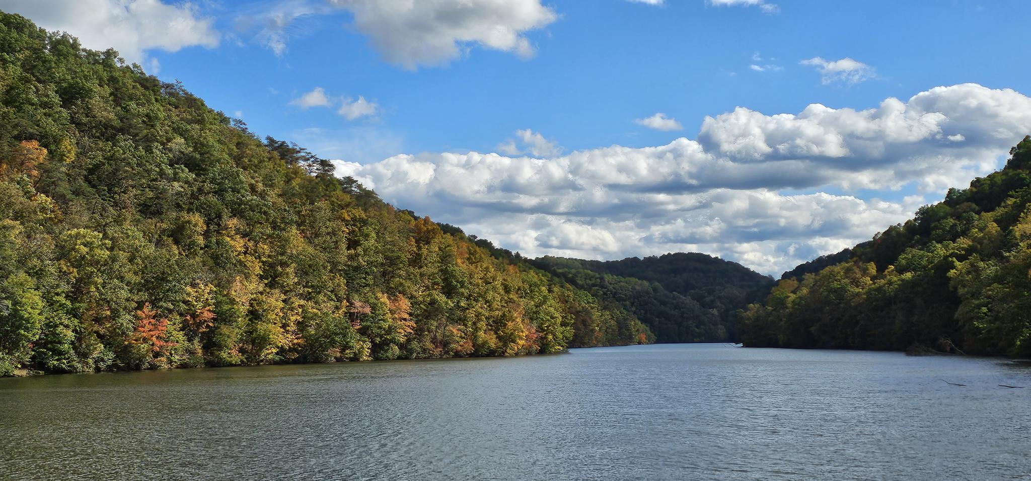 Wide river flowing between tree-covered hills with autumn colors under a cloudy blue sky.