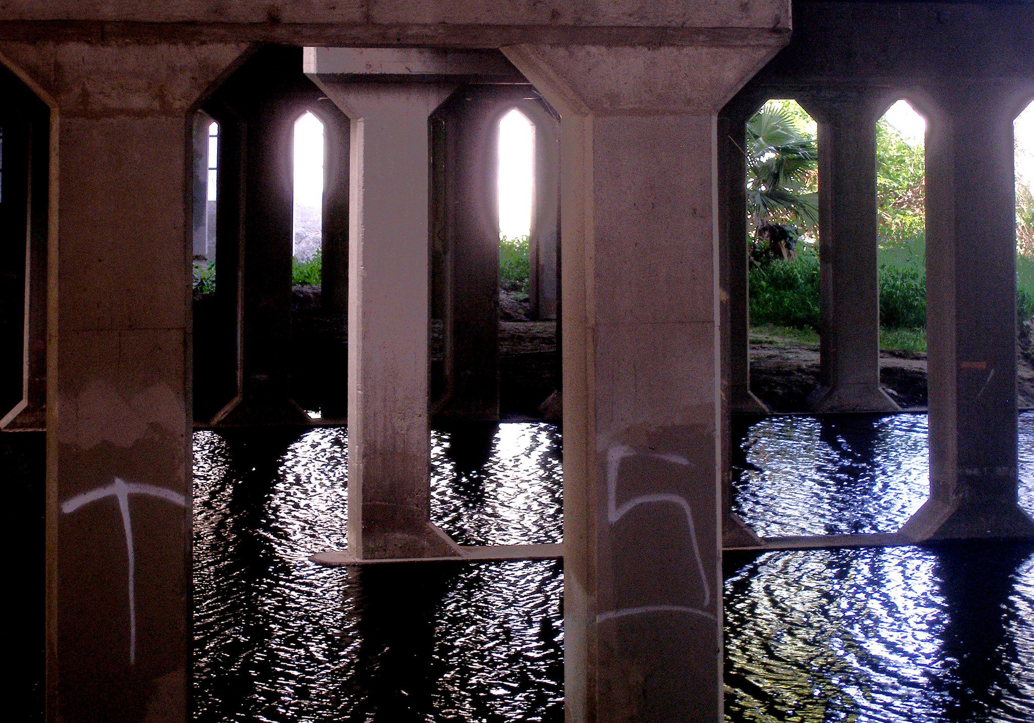 Underneath a bridge, concrete pillars stand in dark, reflective water with bright light.