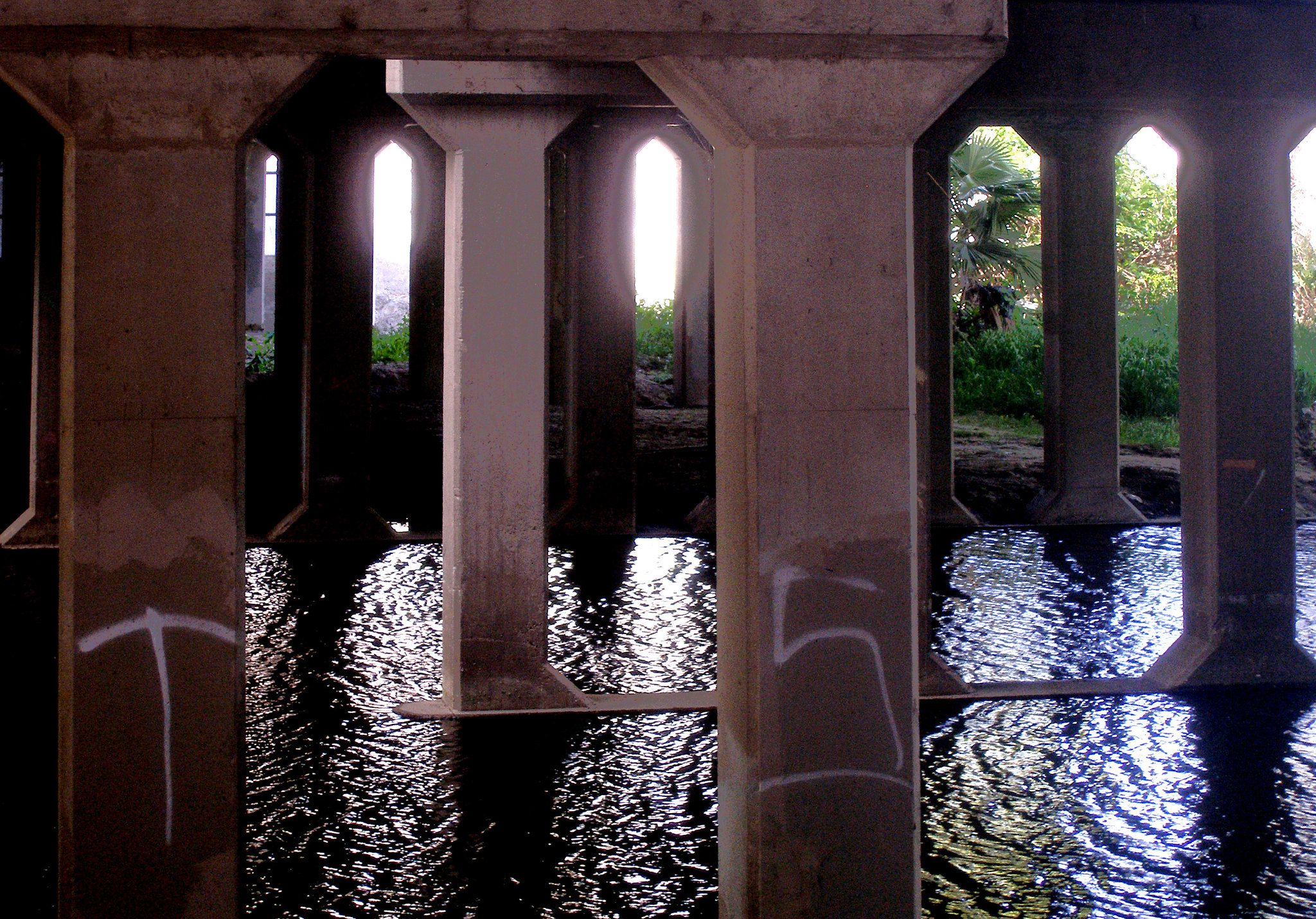 Underneath a bridge, concrete pillars stand in dark, reflective water with bright light. - Sepulveda Basin: Upstream Of Balboa