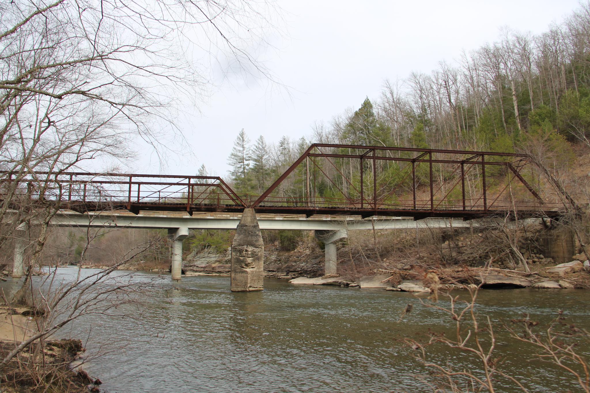 Rusty metal truss bridge spans a river with bare trees under an overcast sky.