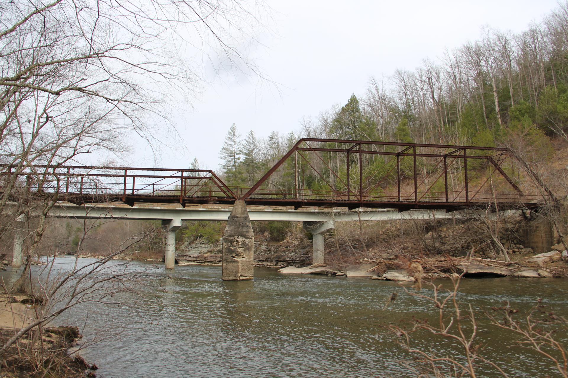 Rusty metal truss bridge spans a river with bare trees under an overcast sky.