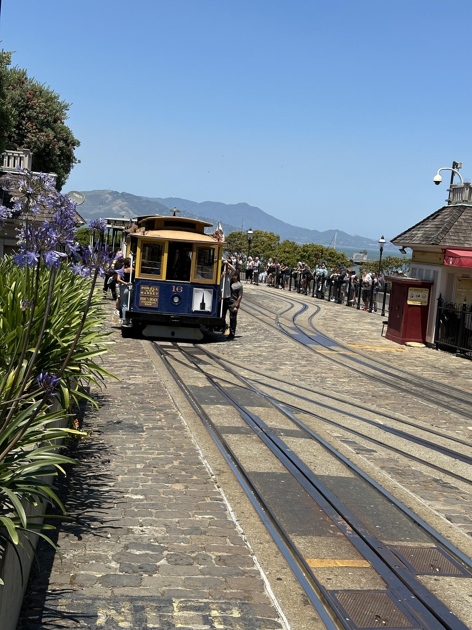 A cable car on tracks, with people waiting and distant hills, sunny day.