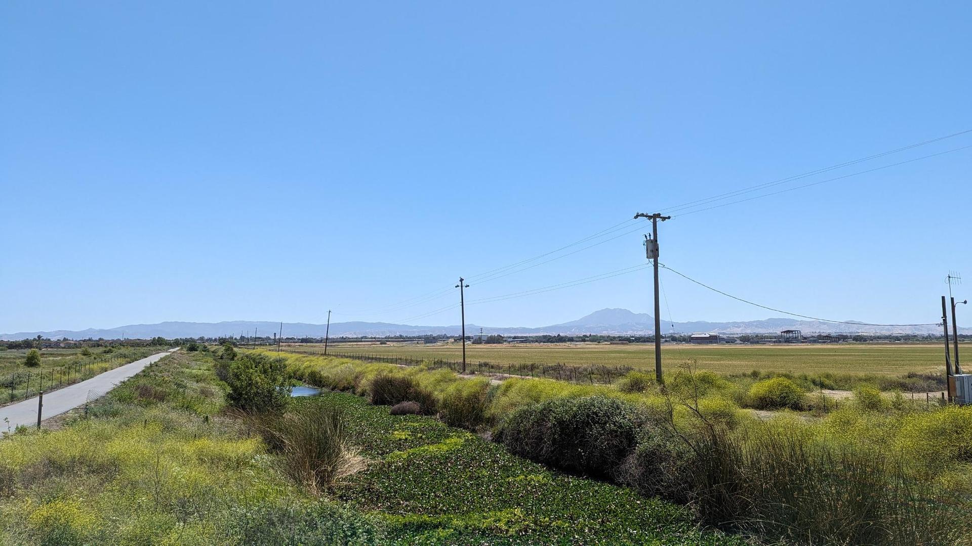 Rural landscape with a dirt road, canal, fields, and distant mountains under clear skies.