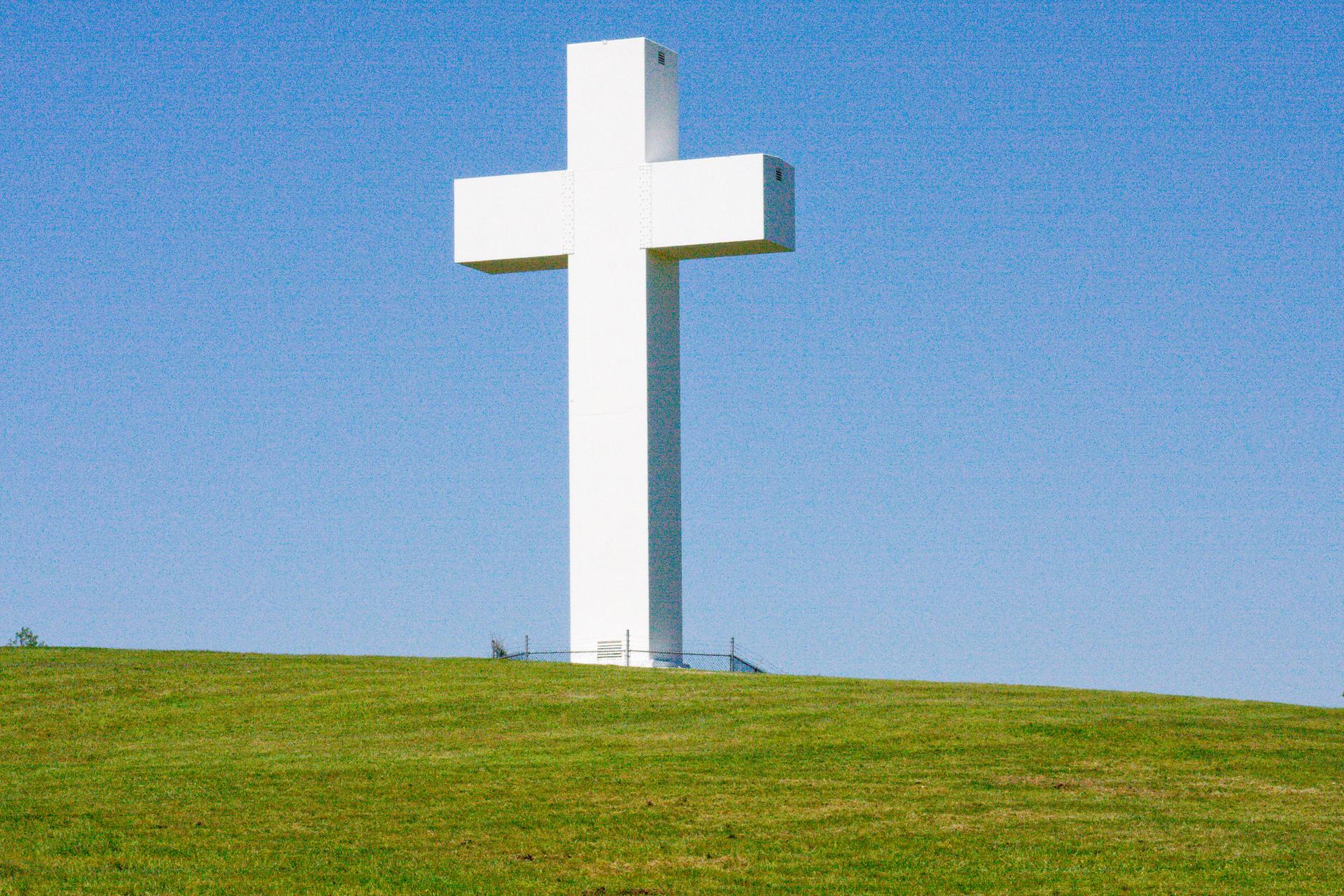 A large white cross stands on a green grassy hill under a clear blue sky.
