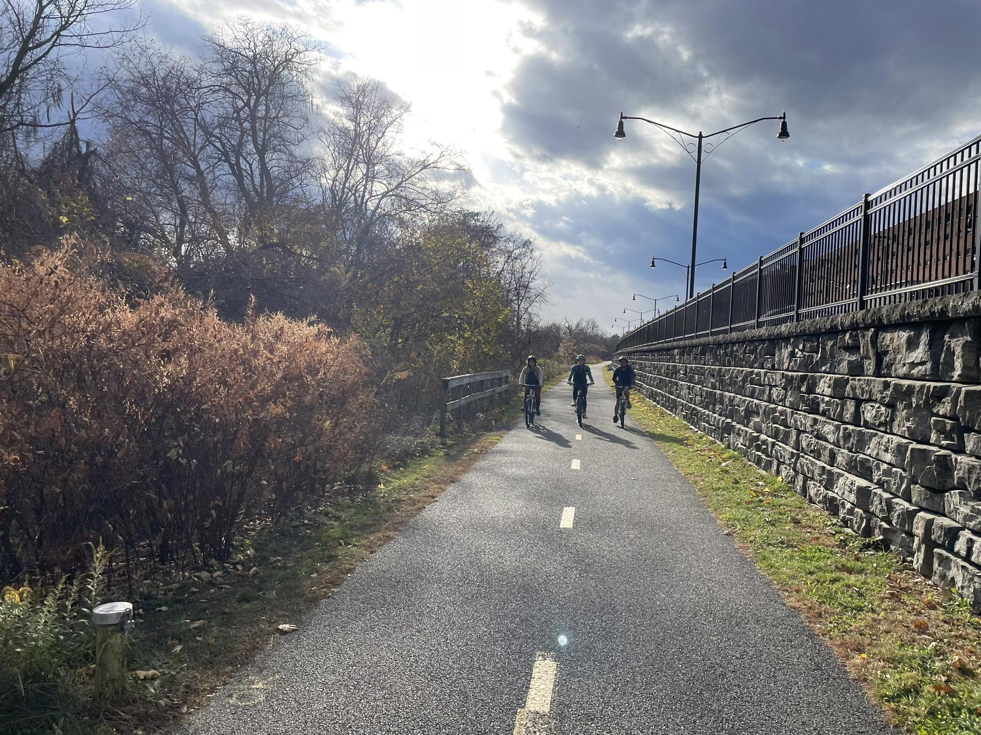 Three people ride bicycles on a paved path next to a stone wall.