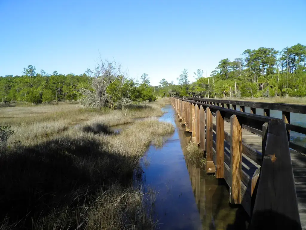 Long wooden boardwalk extends over calm water and marshland under a clear blue sky.