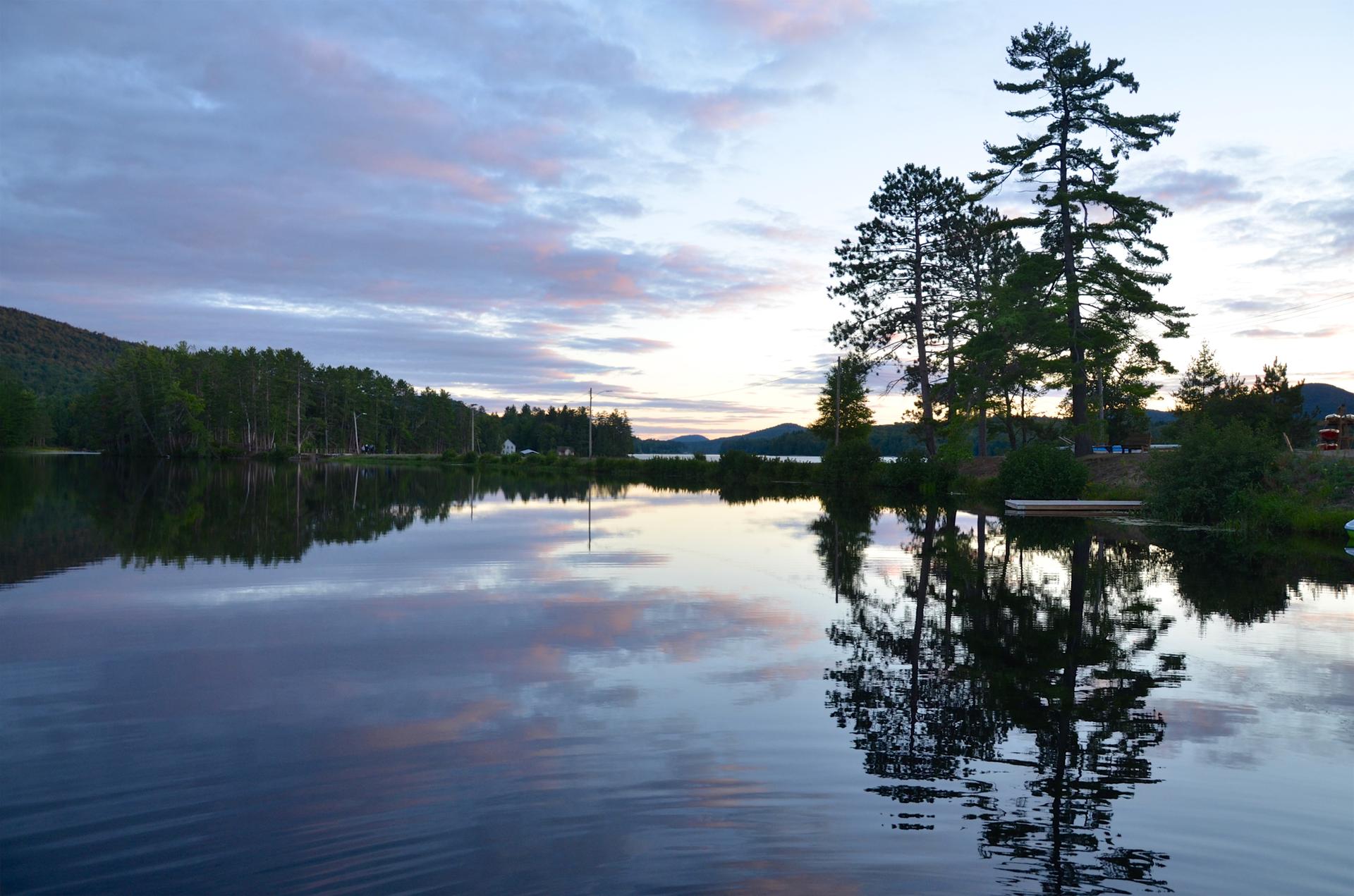 Still lake reflecting tall pine trees and a colorful cloudy sky at dusk.