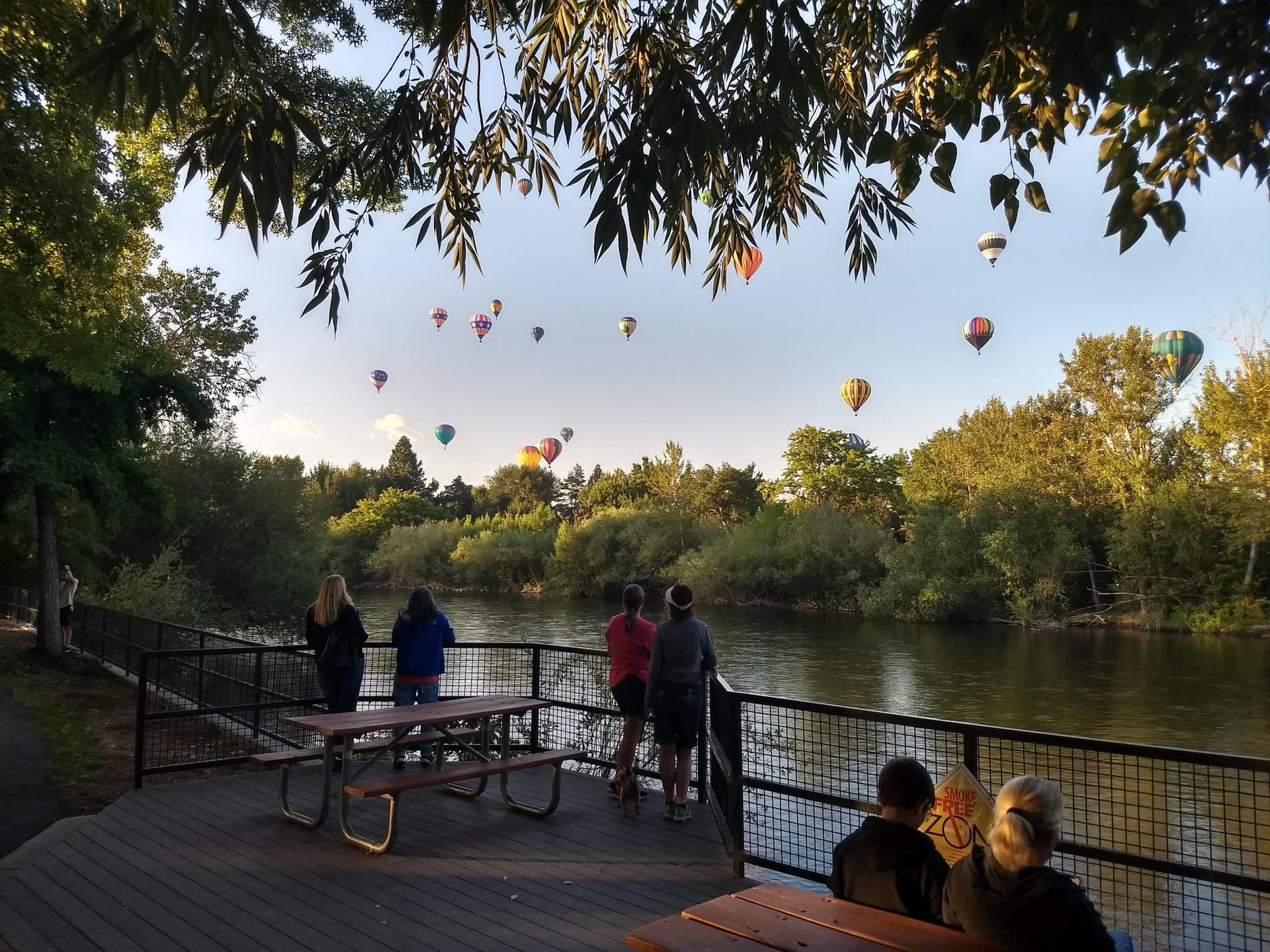 People watch colorful hot air balloons ascend over a river at sunset.