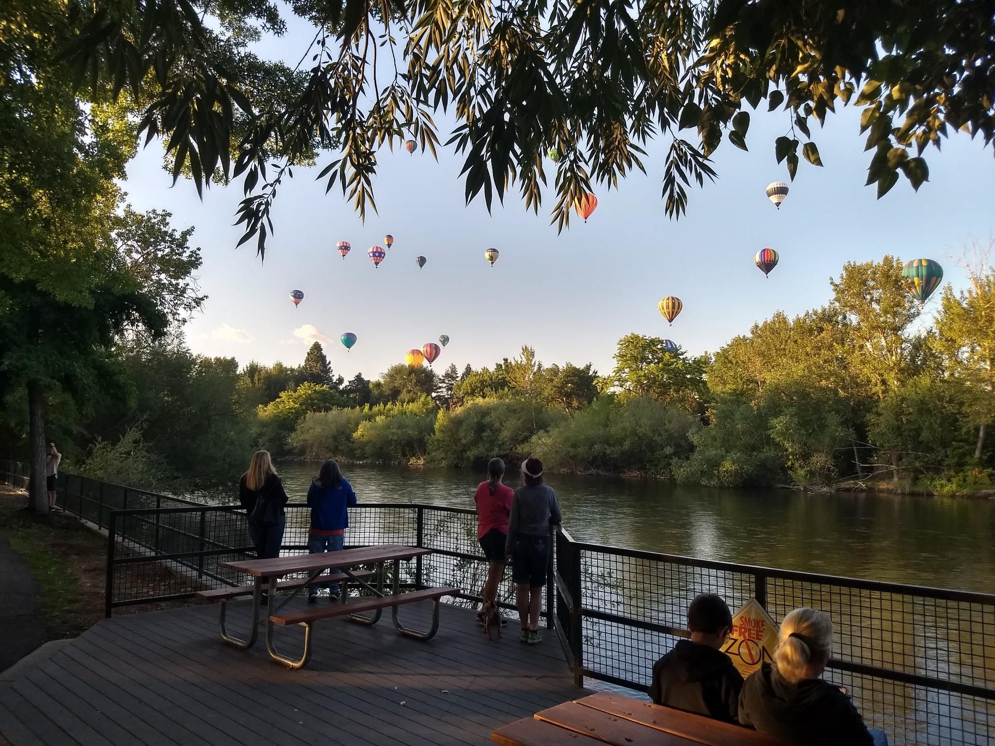People watch colorful hot air balloons ascend over a river at sunset. - Boise River Greenbelt – Boise river view loop from Boise