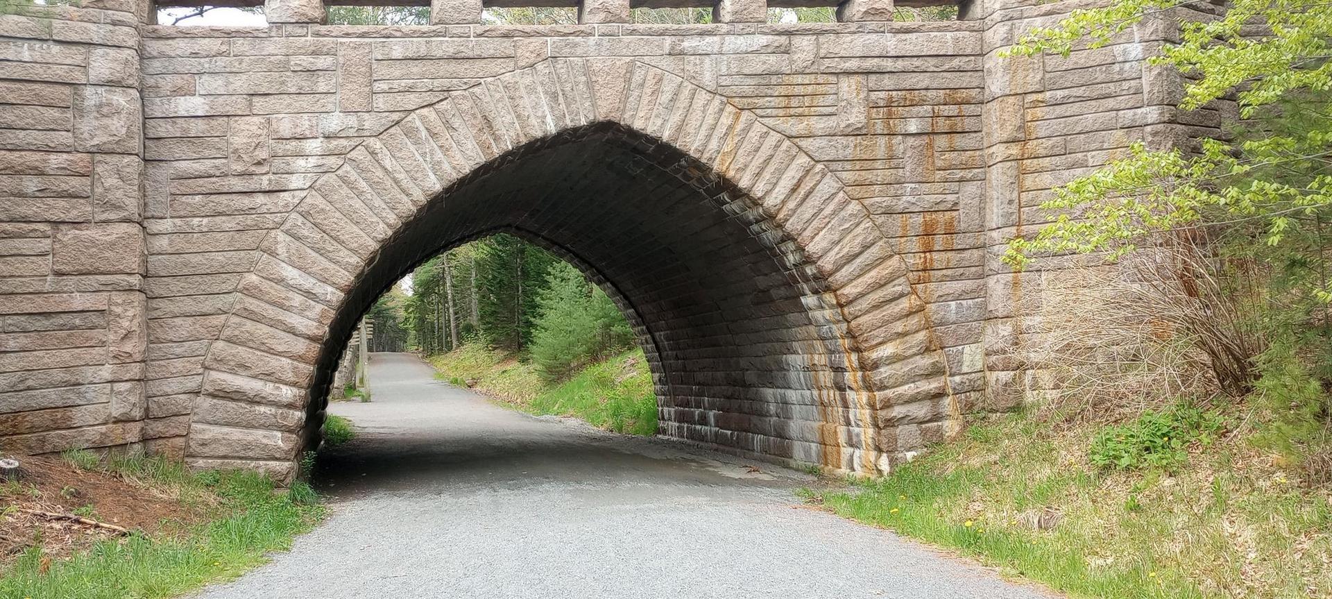 Stone archway of a bridge over a gravel path, surrounded by green foliage.