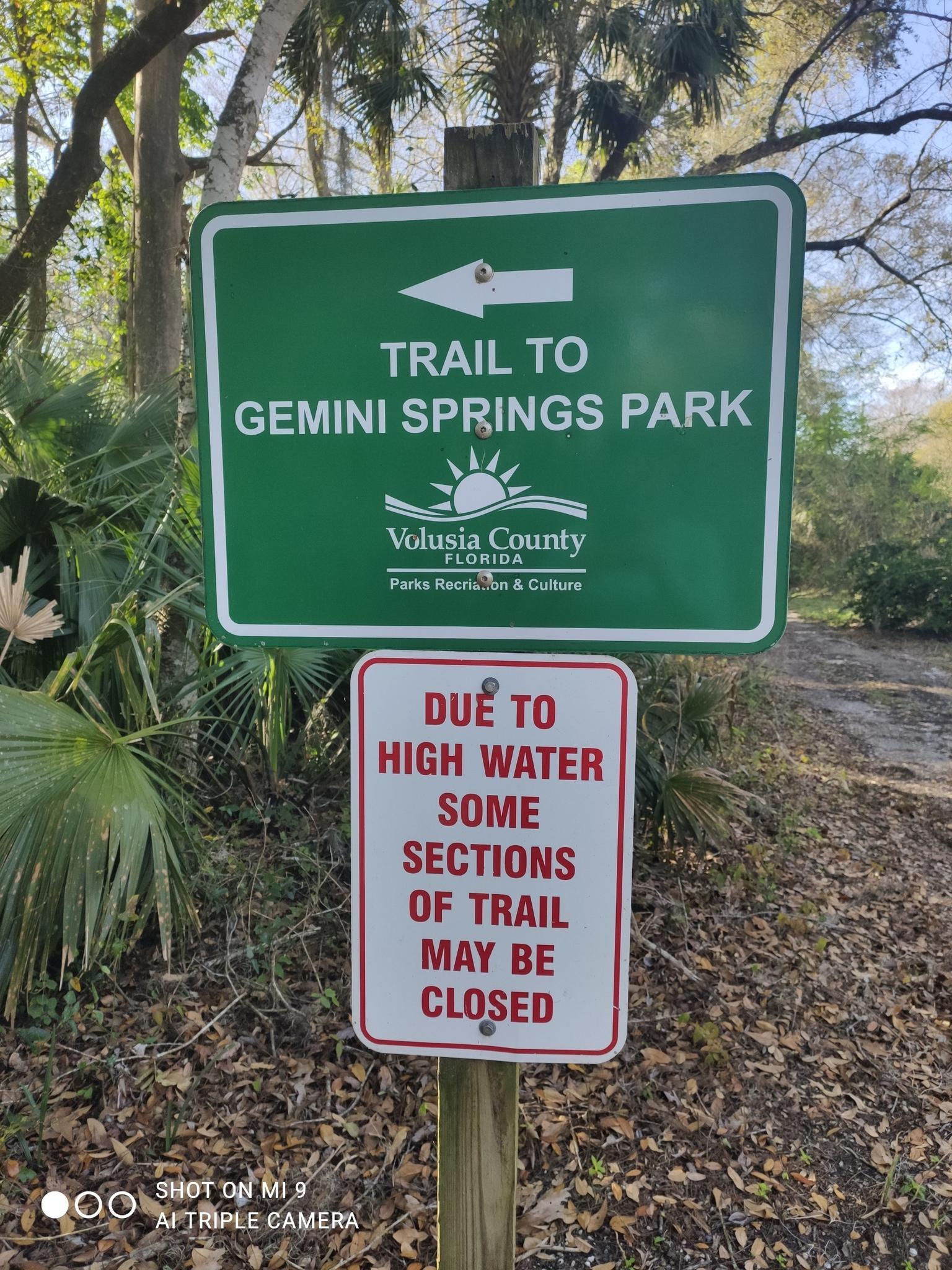 Two trail signs, one warning of high water, stand in a sunny, leafy forest.