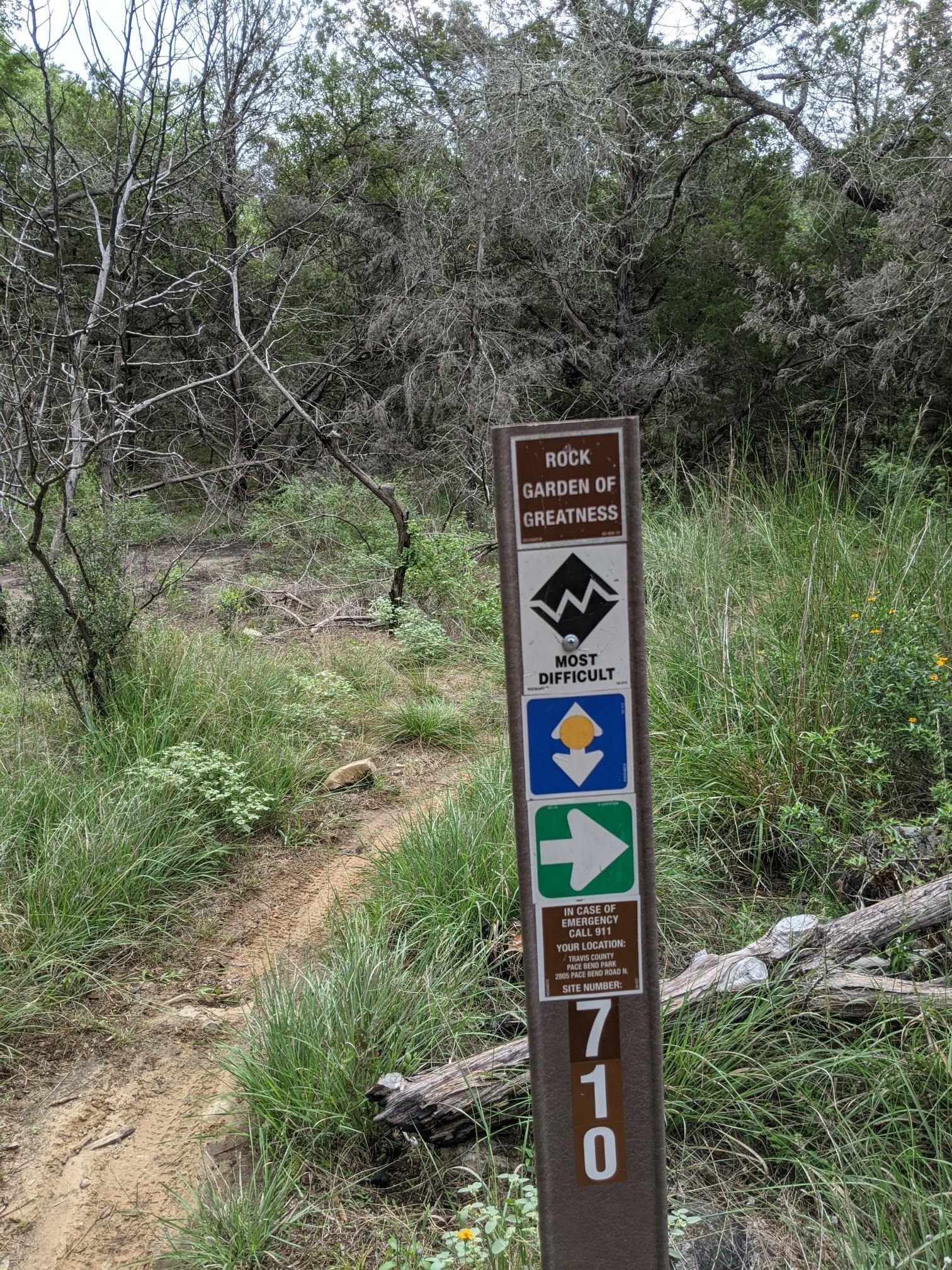 A brown trail sign stands beside a dirt path in a wooded, grassy area.