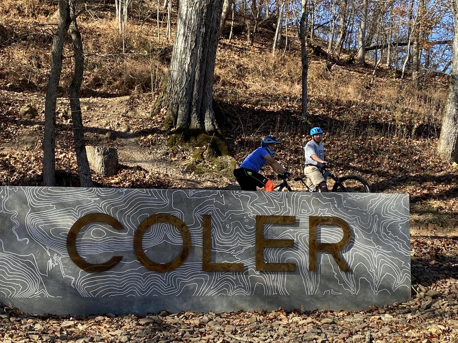 Two mountain bikers ride past a large stone sign in a sunny forest.