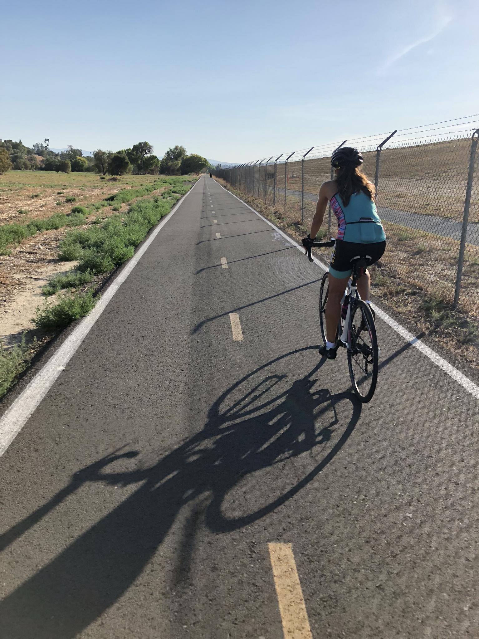 A person cycles on a paved path with a long shadow on a sunny day.