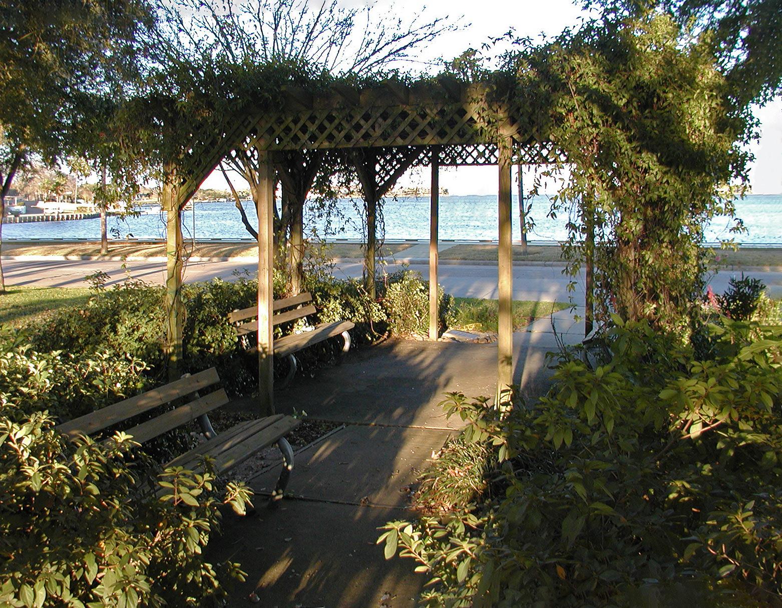 Shaded wooden pergola with benches overlooking calm water on a sunny day.