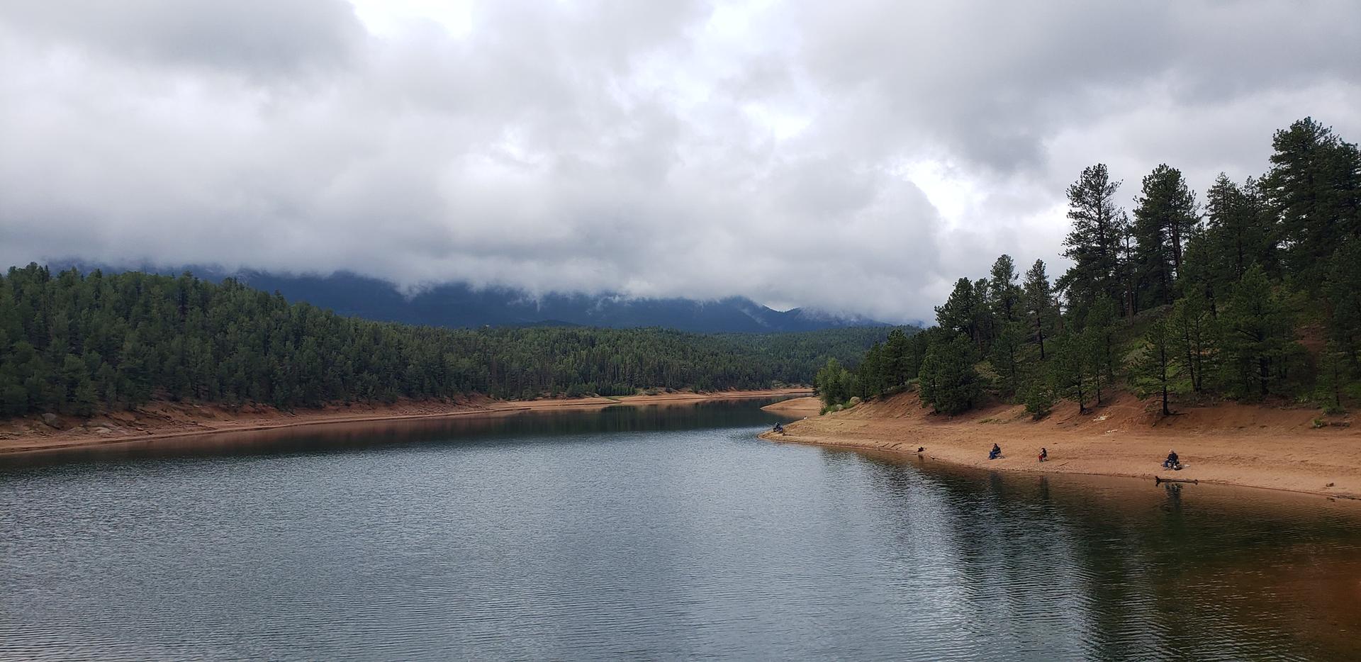 A cloudy lake with pine forests, sandy banks, and people on the shore.