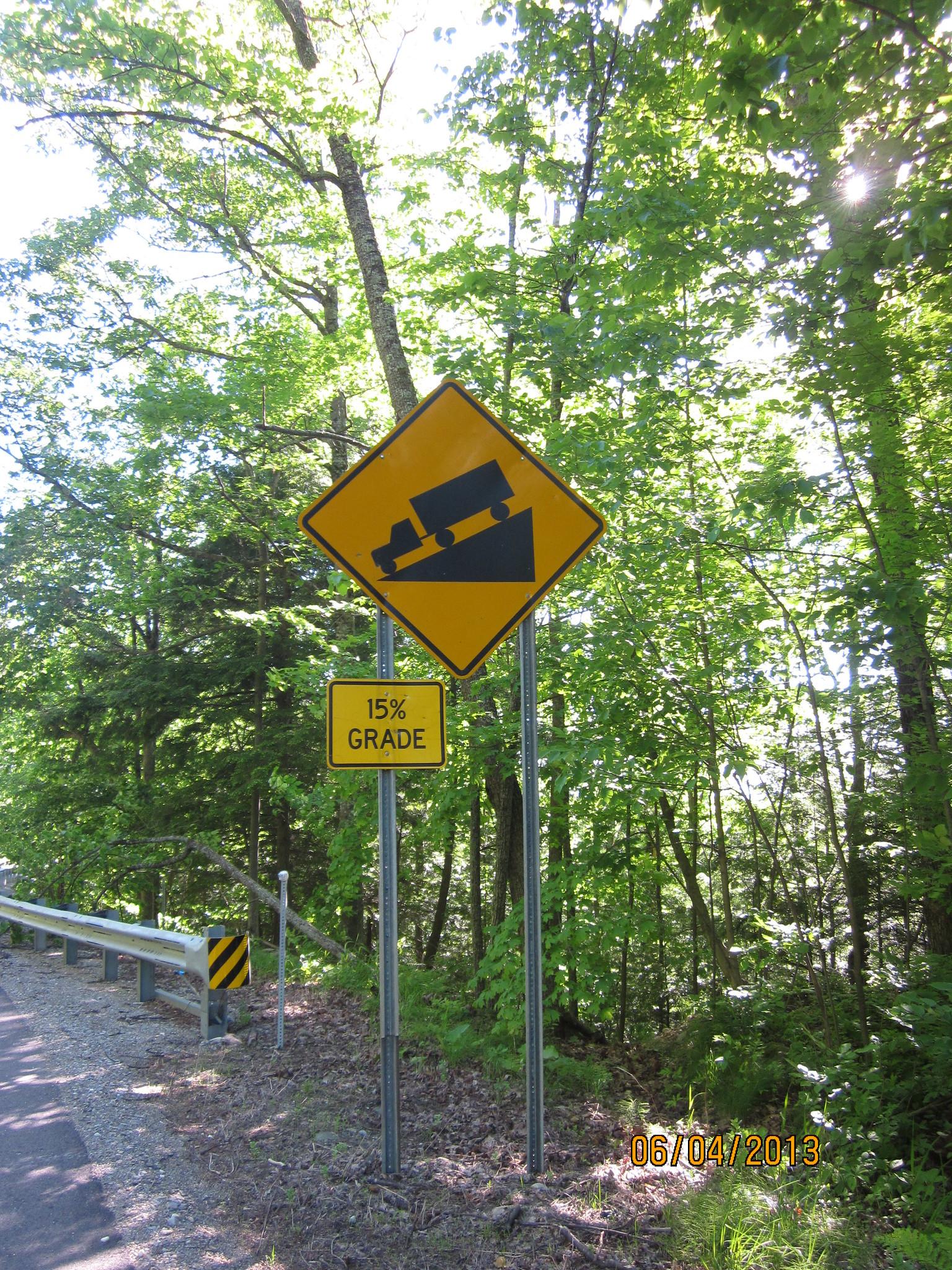 A steep grade warning sign on a sunny forested road with a guardrail.