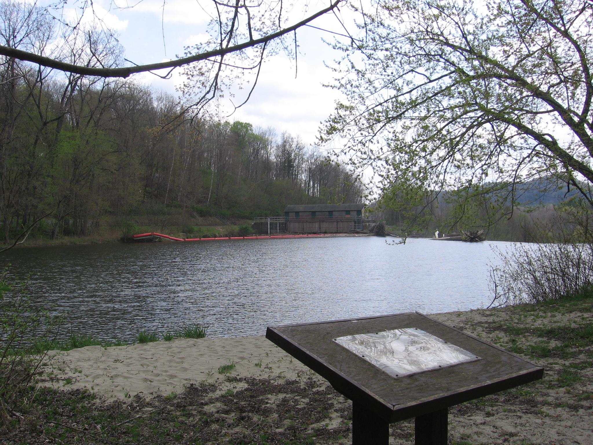 A lake with wooded hills and a distant building, an information sign in foreground.
