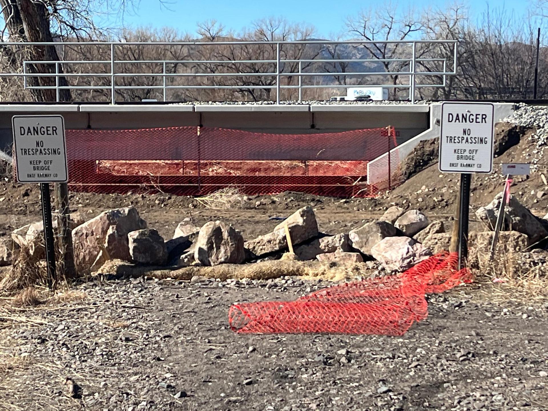 Bridge underpass with danger signs, red fencing, and rocks on a sunny day.