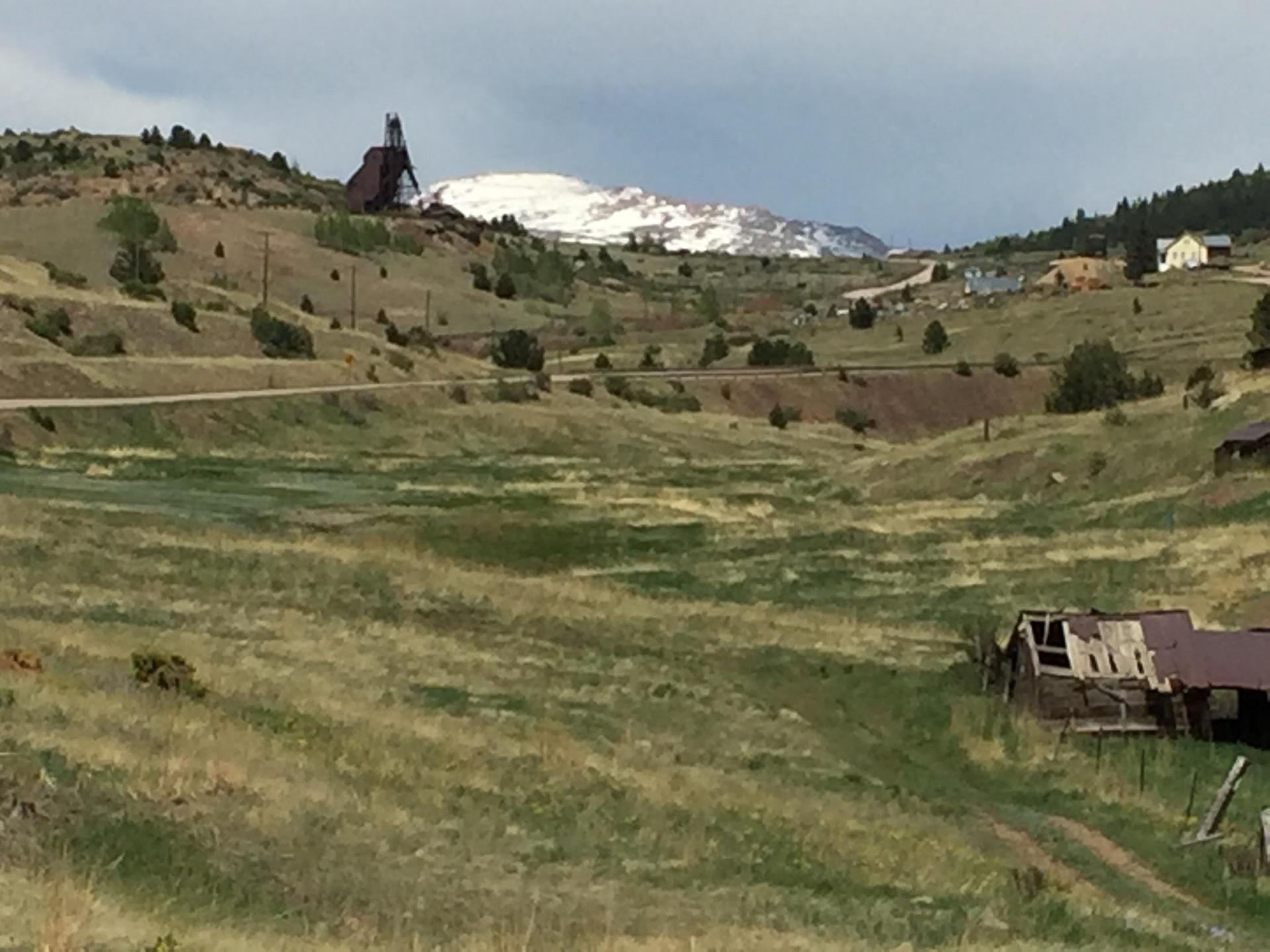 Hilly, grassy landscape with an old building, road, and snow-capped mountain under cloudy sky.