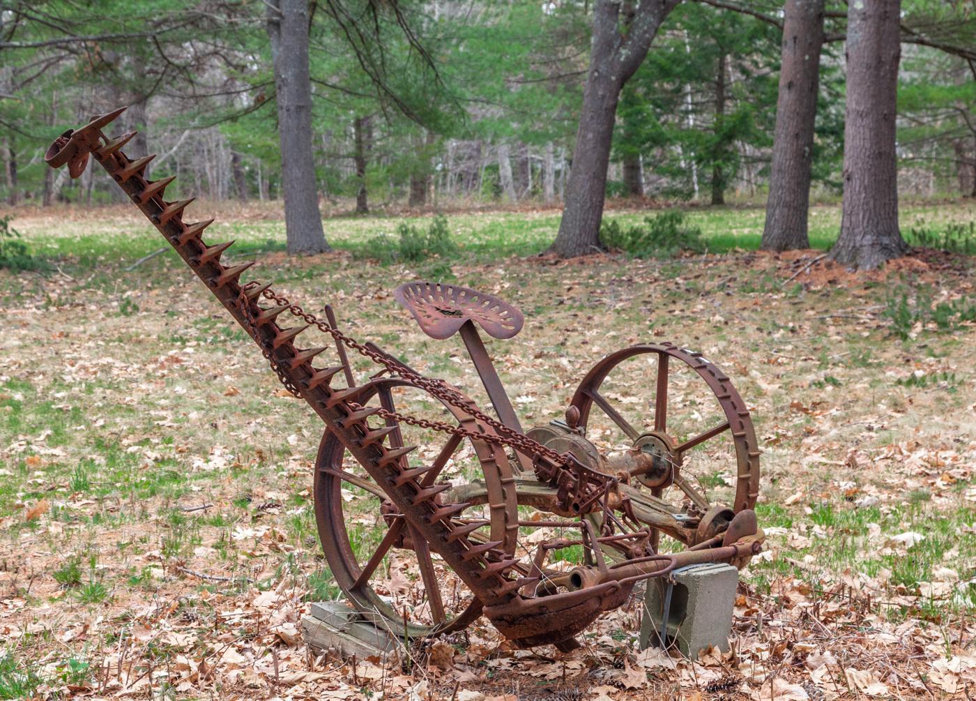 Rusty antique farm machinery sits on leafy ground with trees in the background.