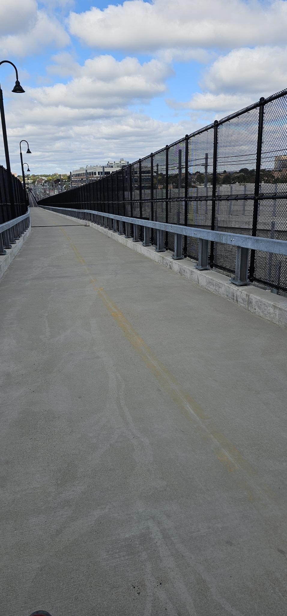Empty concrete pathway with a chain-link fence under a cloudy sky.