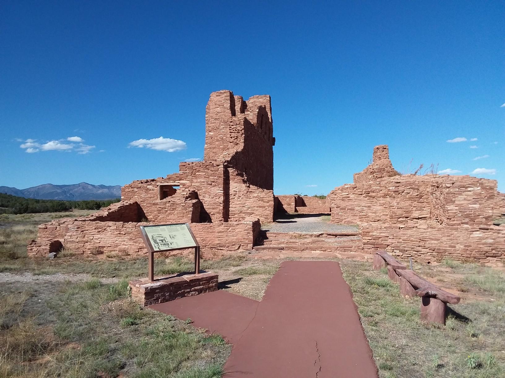 Ancient red stone ruins with a paved path and informational sign under blue sky.