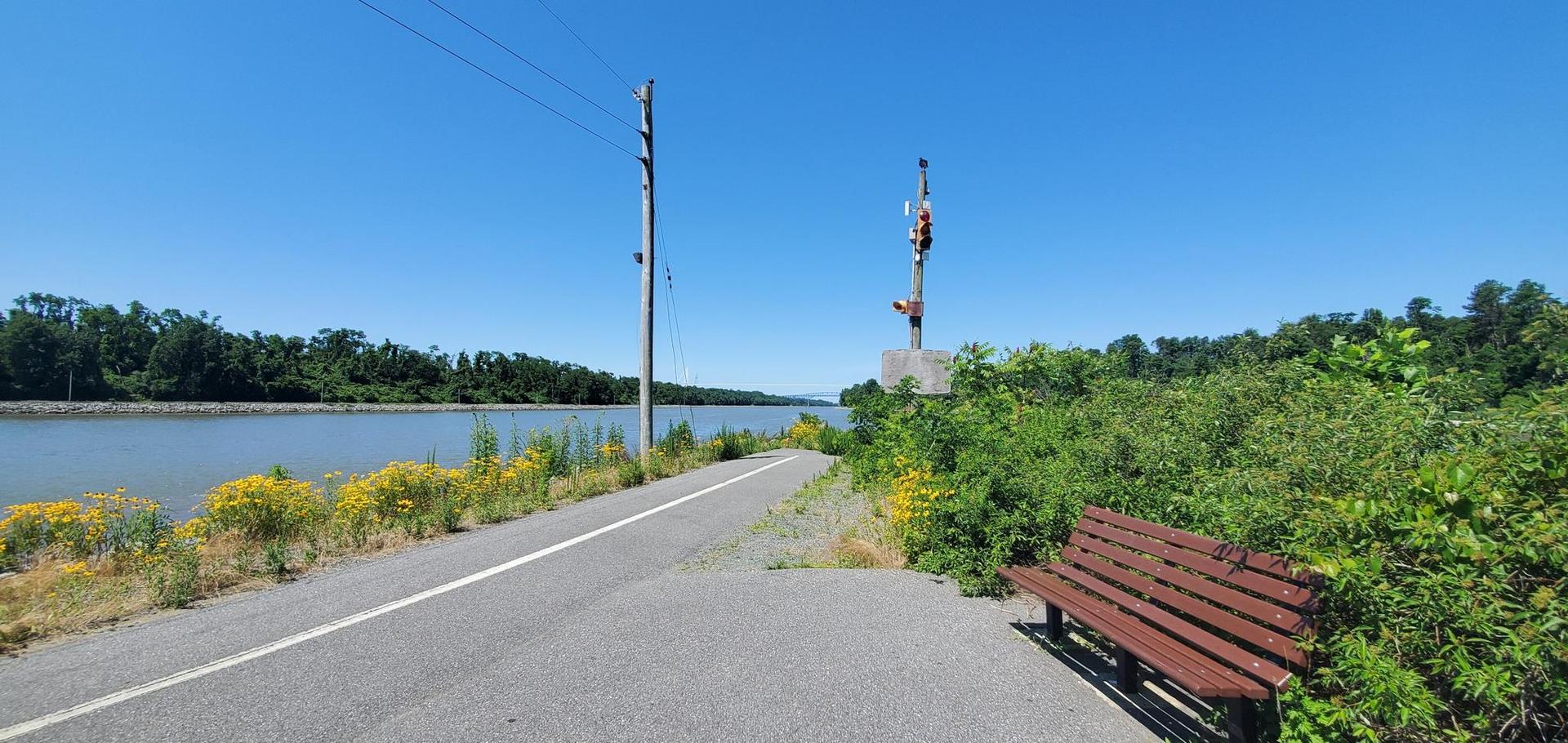 Paved path alongside a river with a bench under a clear blue sky.