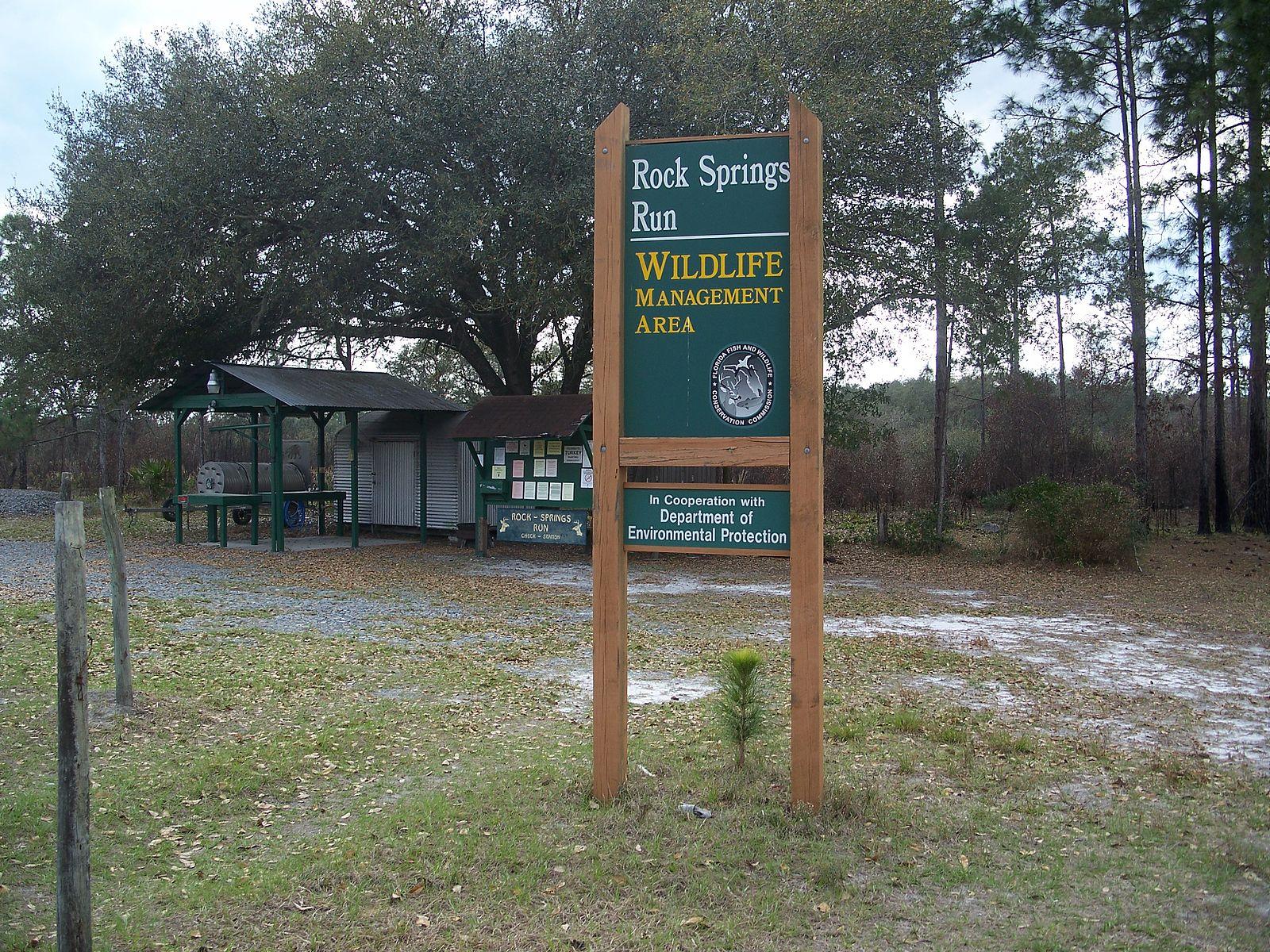 A large green sign stands in a grassy area near trees and a shelter.