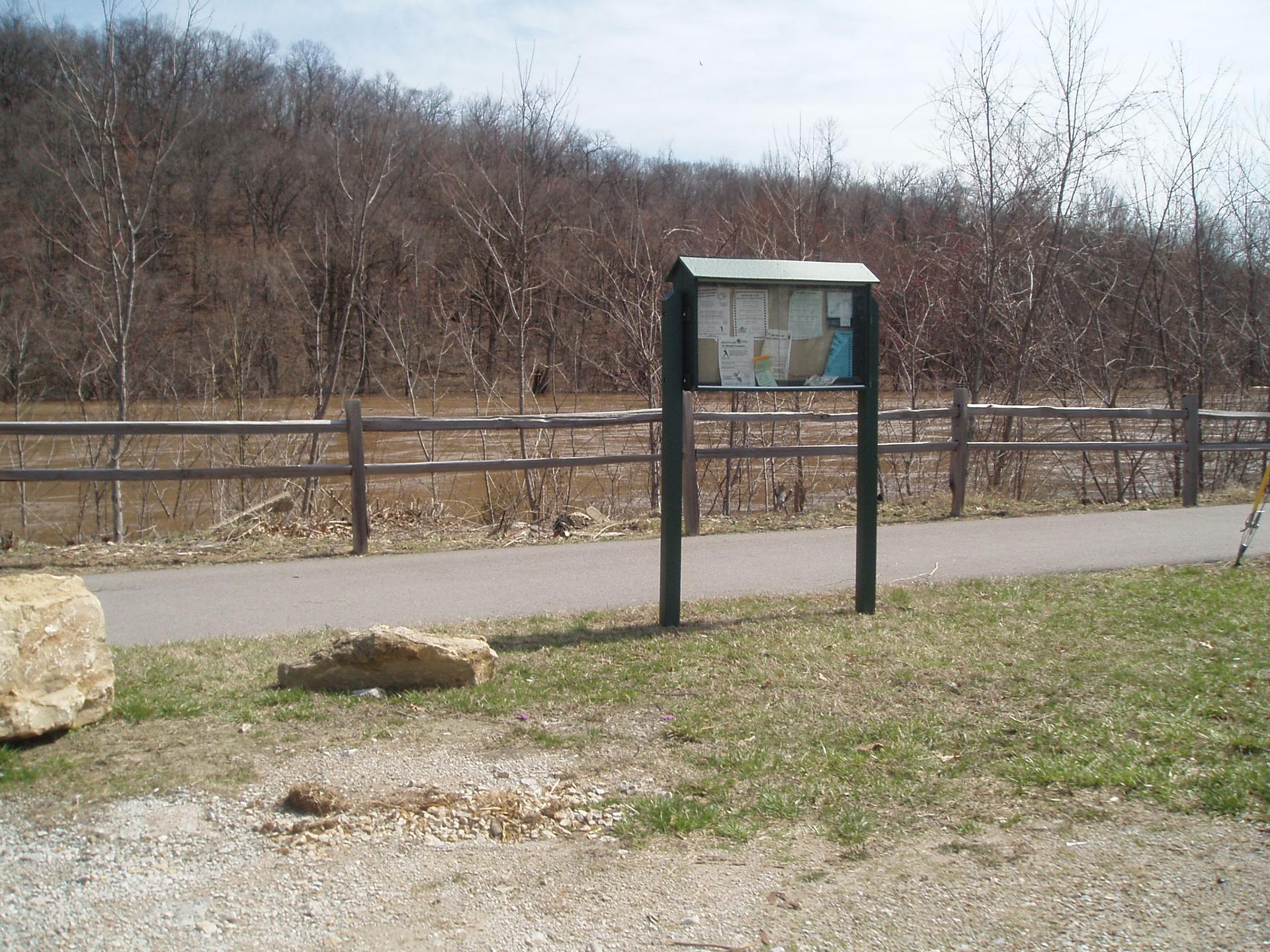 An information board stands beside a paved path and wooden fence.