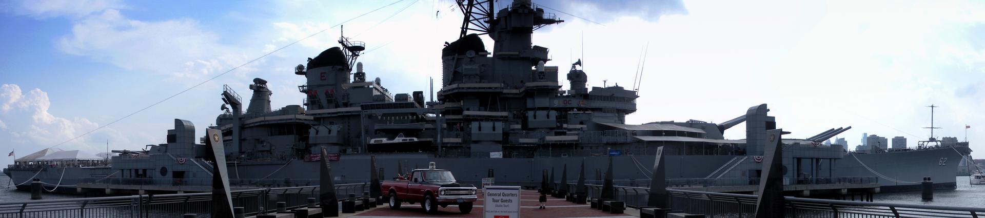 A large battleship docked under a bright, overcast sky, with a red truck nearby.