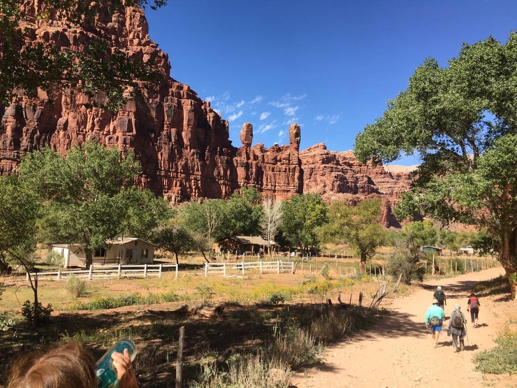 Hikers on a dirt path, red rock cliffs and green trees under a clear sky.