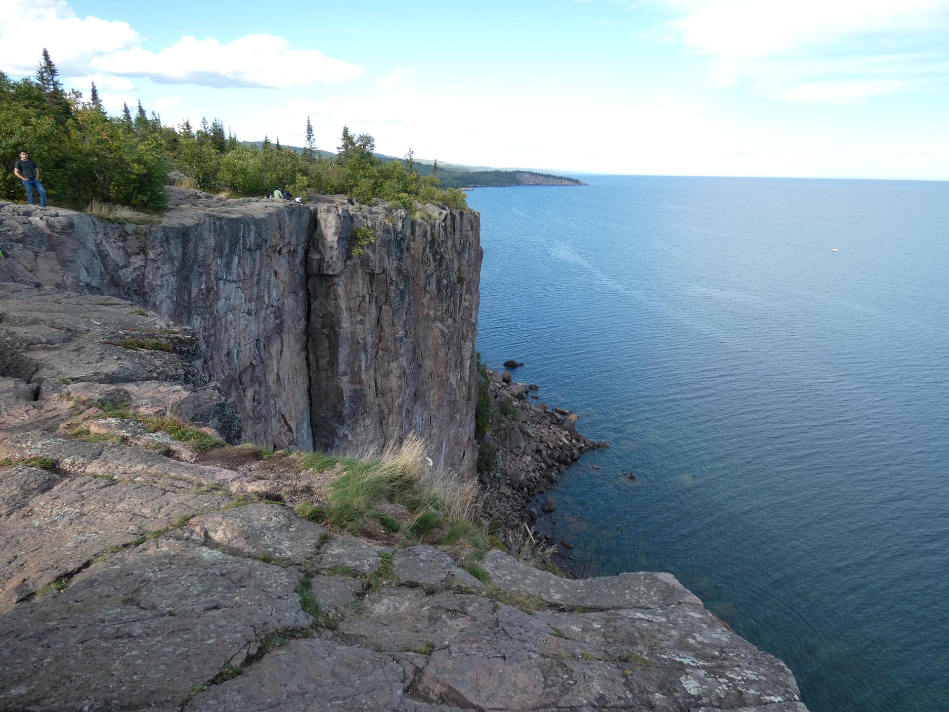 Steep rocky cliff overlooking a vast blue lake with a person standing.