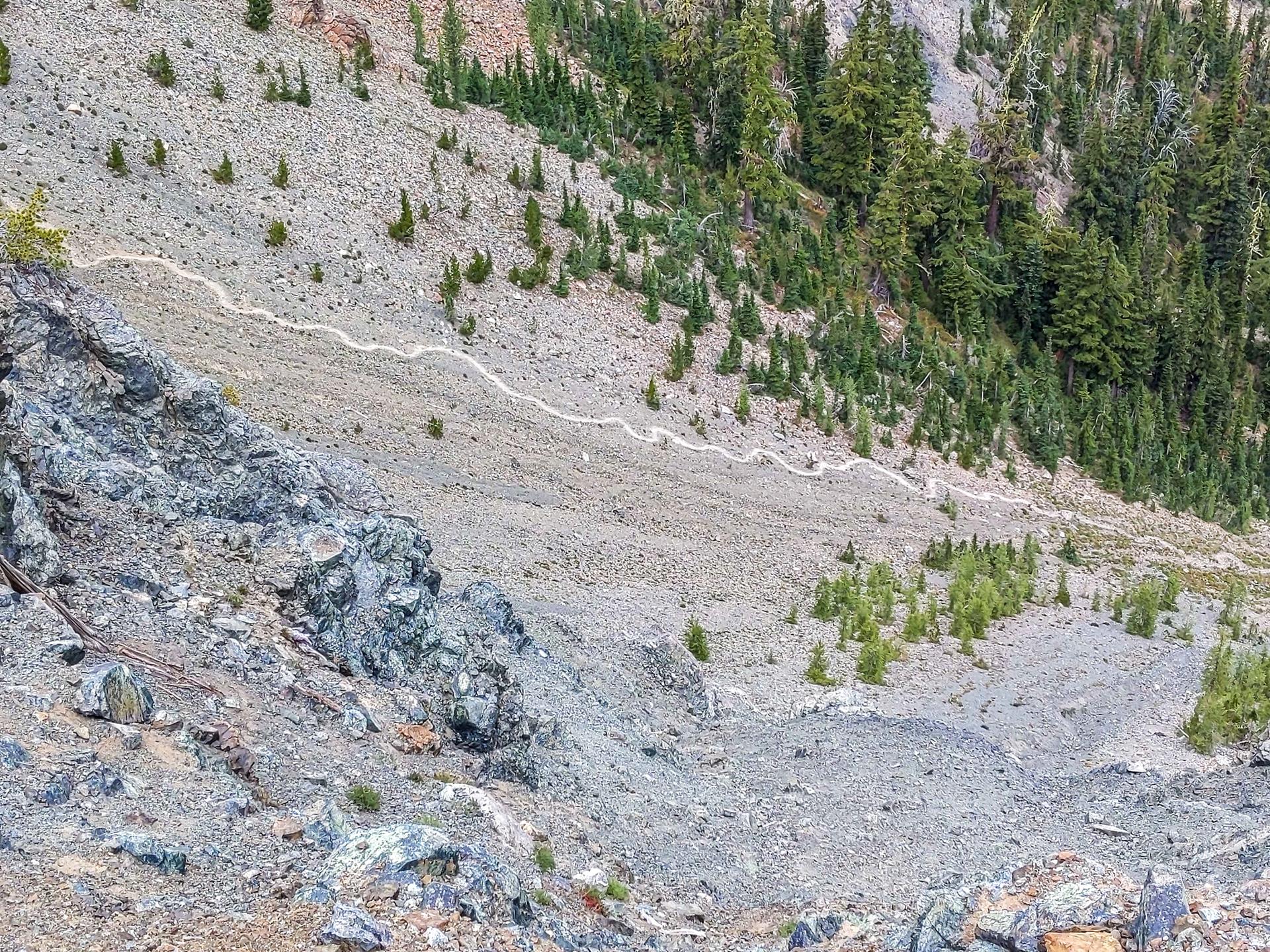 Steep, rocky mountainside with sparse evergreen trees and a winding path under diffused light.