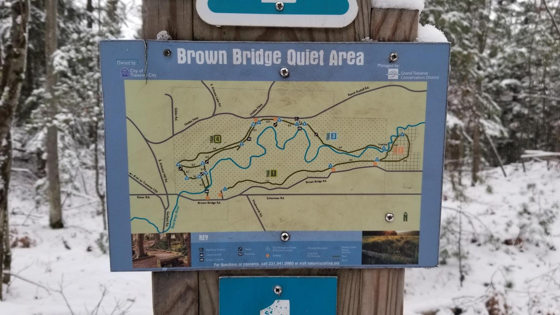 A trail map sign stands in a snowy forest with tall, bare trees.