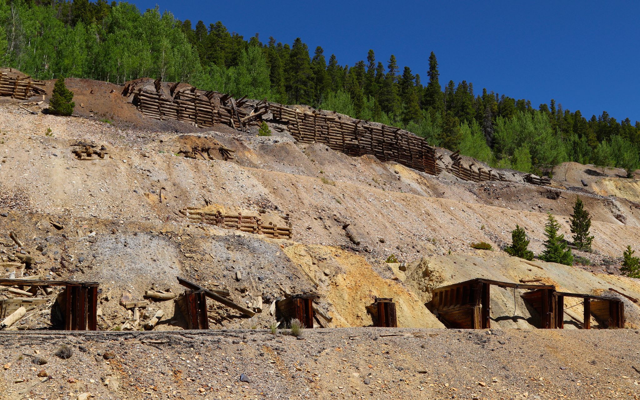 Terraced hillside with wooden support structures and small mine entrances under a blue sky. - Nugget Gulch – Matchless Mine loop from Leadville
