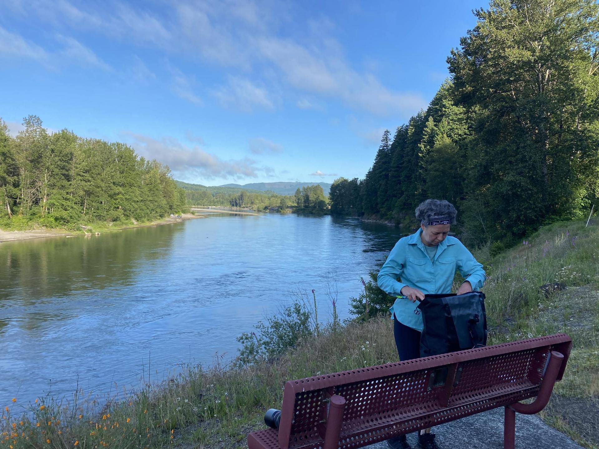 Woman in blue shirt looking into a bag by a river on a sunny day.