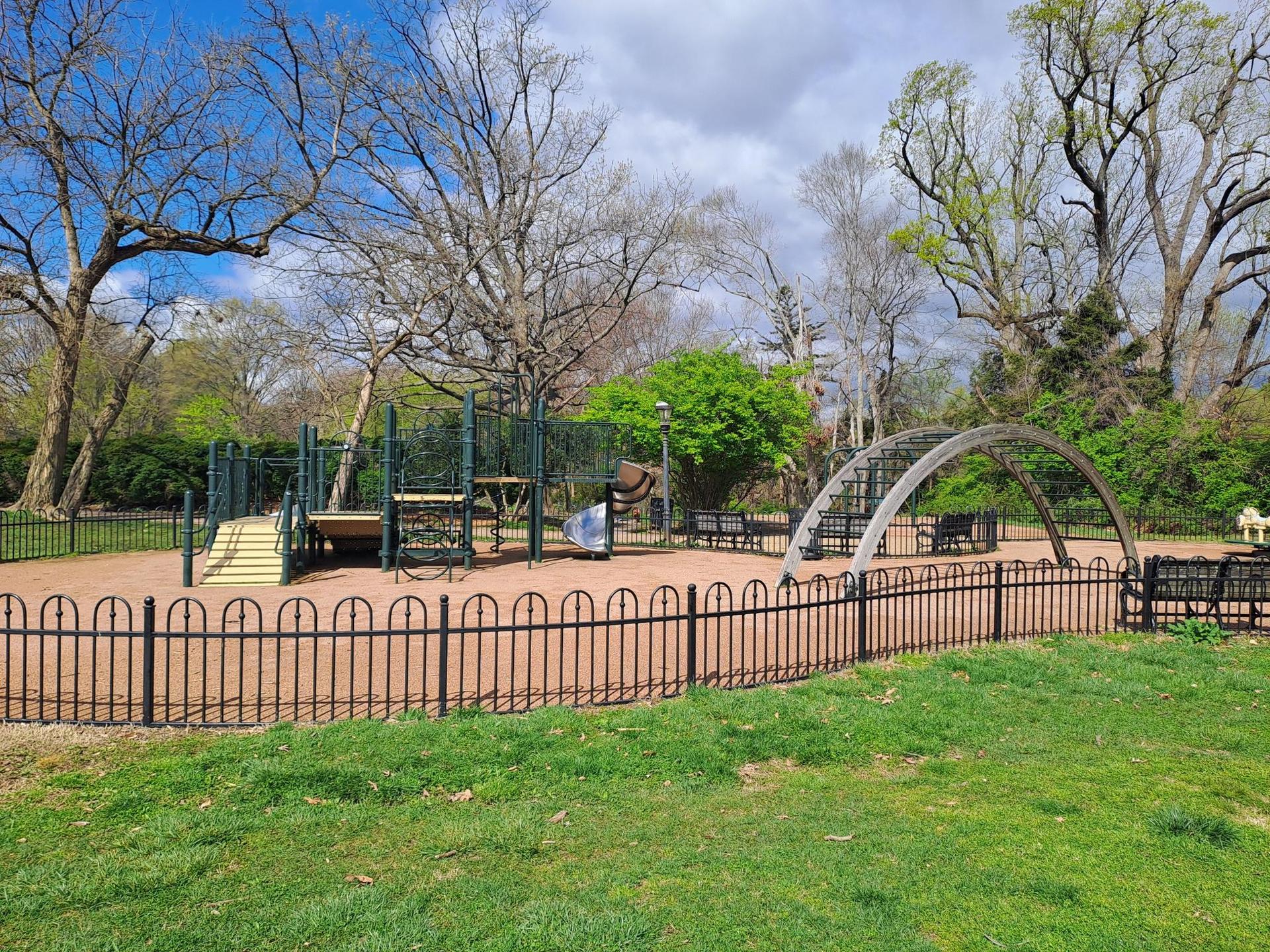 An empty playground with metal structures and a fence, surrounded by trees.