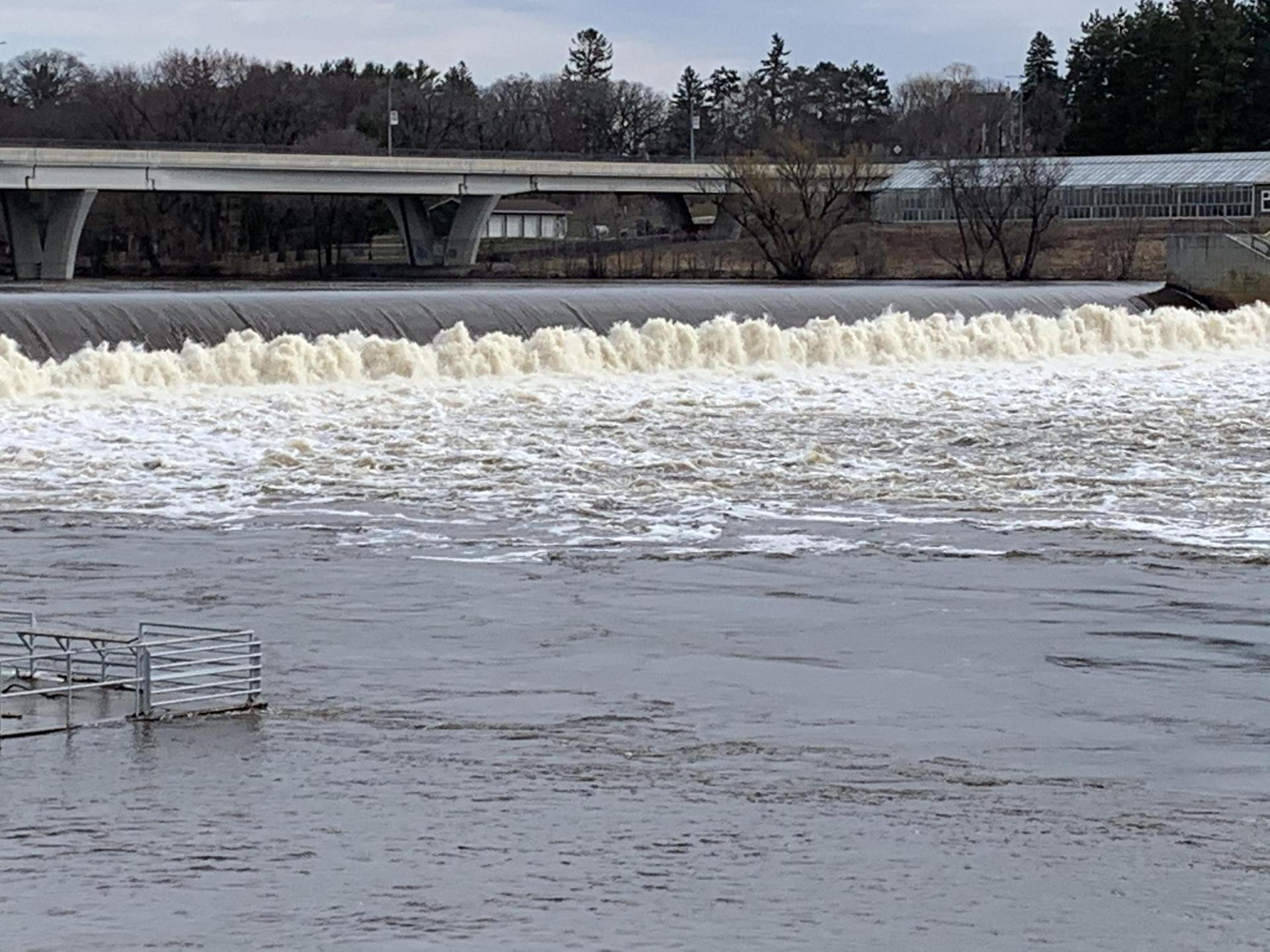 Foaming river dam with a bridge and bare trees under an overcast sky.