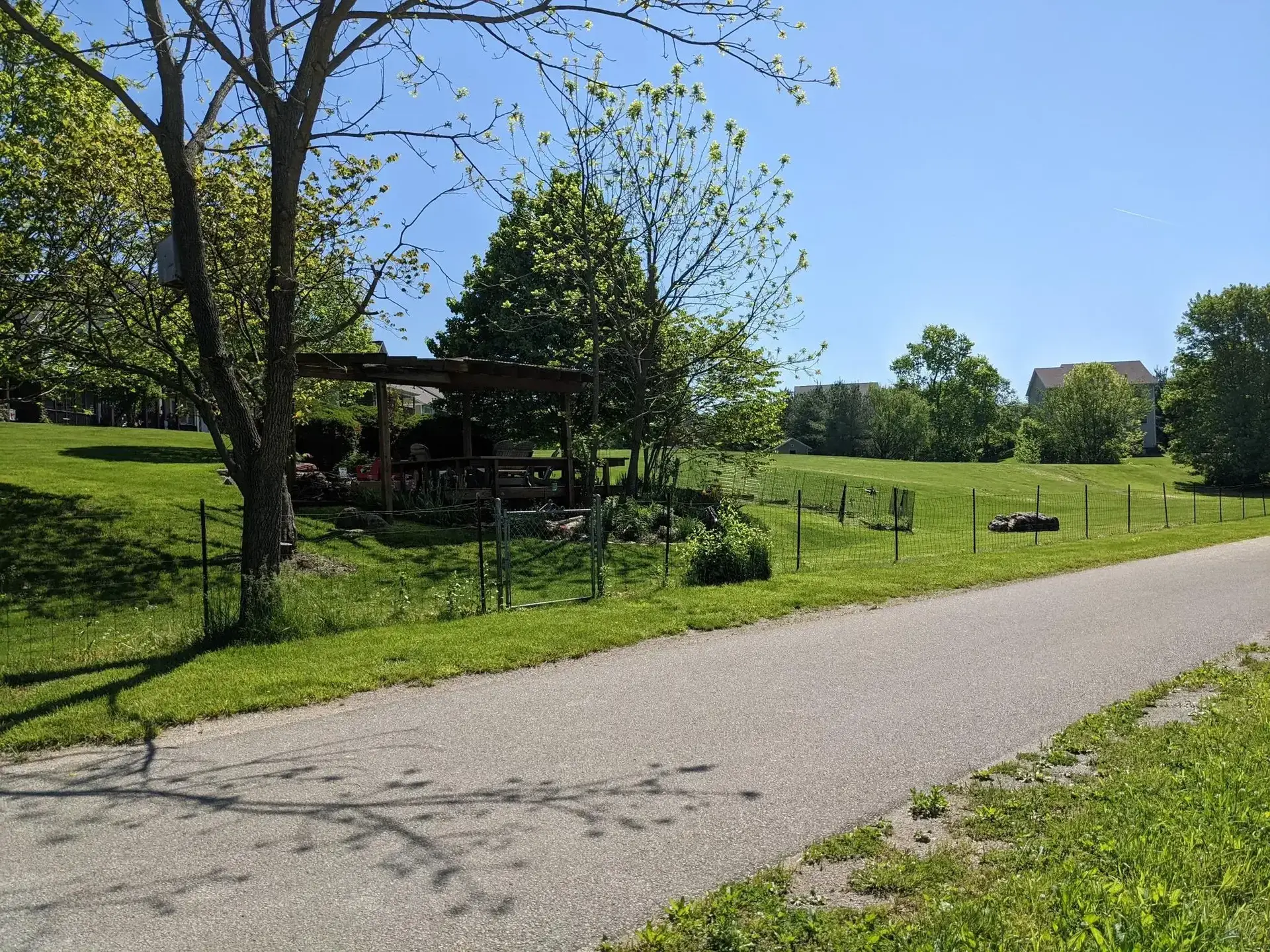 A paved path runs alongside a sunny green lawn with trees and a fence.