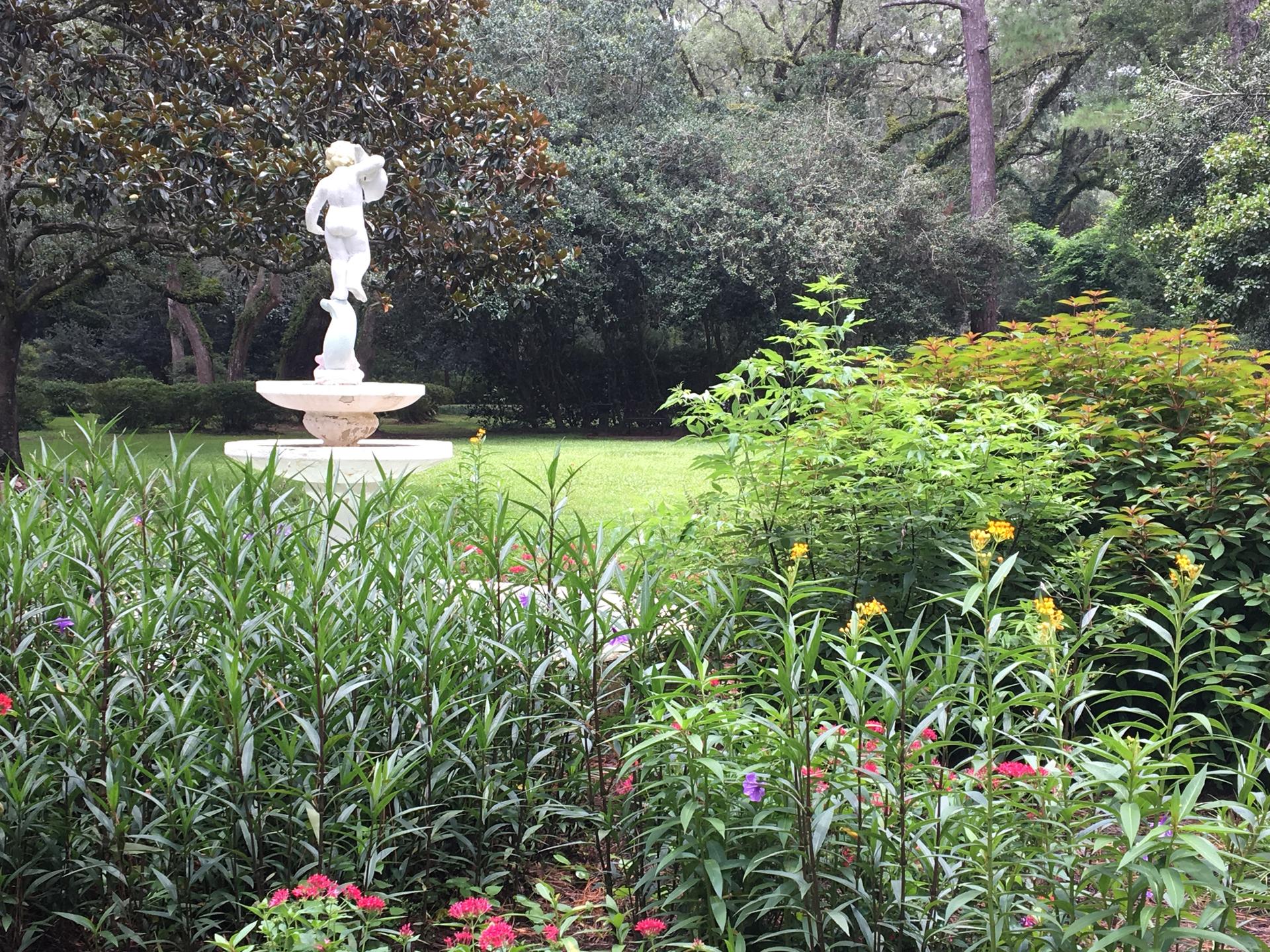 White statue fountain in a lush green garden with colorful flowers under bright light.
