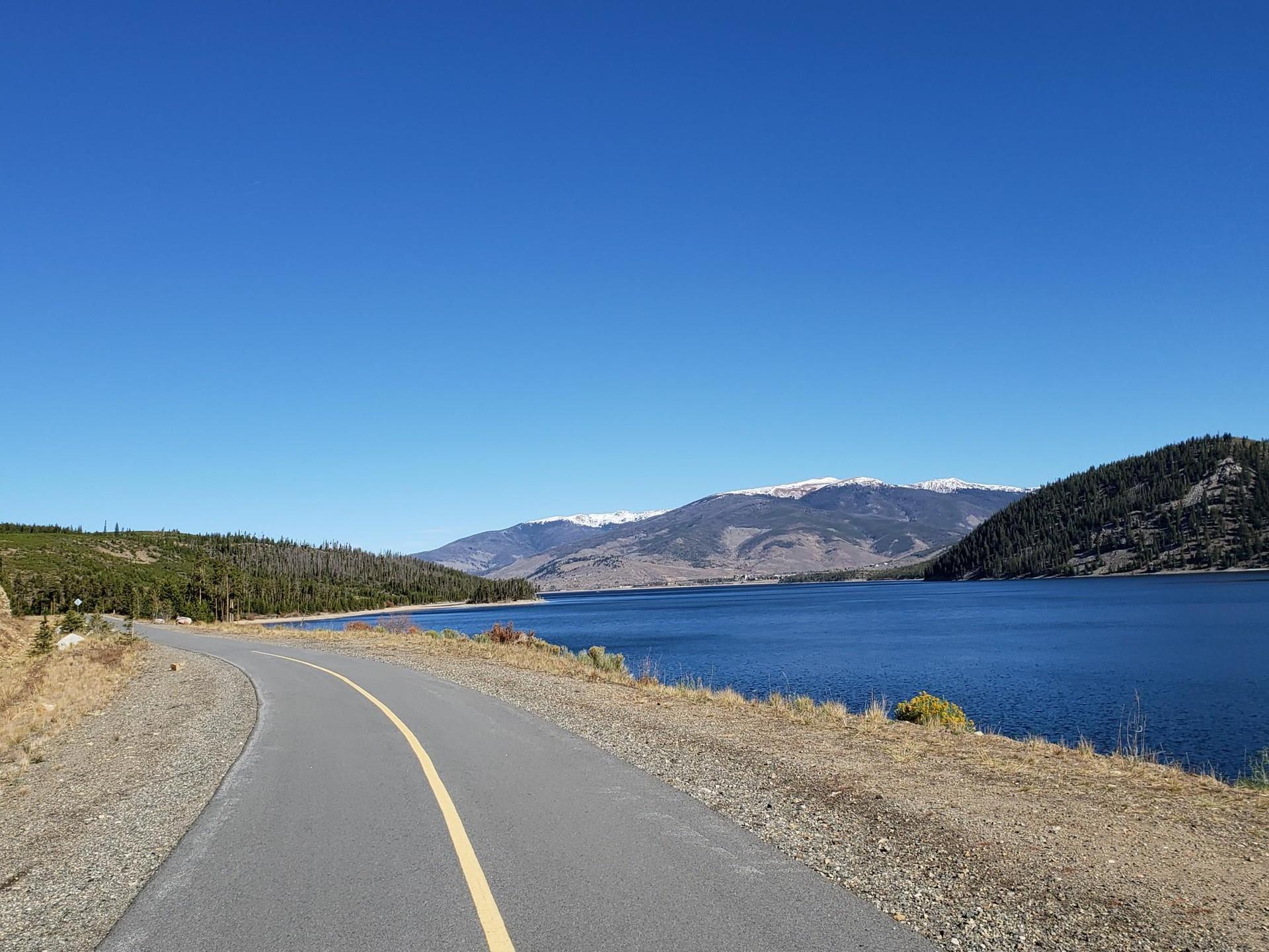 Paved road alongside a large blue lake, forested hills, and snow-capped mountains.